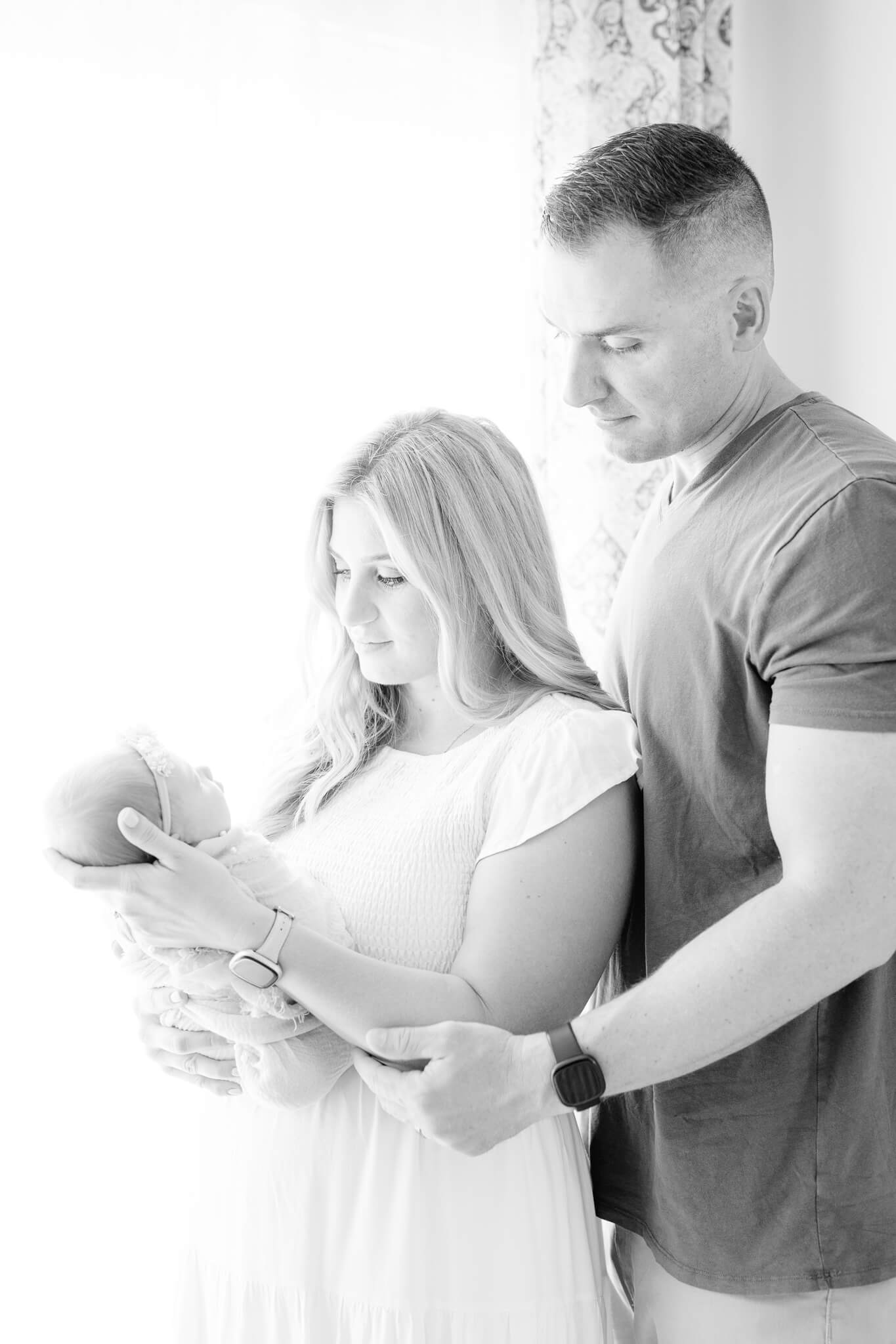 mom and dad holding their newborn baby girl in front of a curtained window Southern Maryland Newborn Photographer