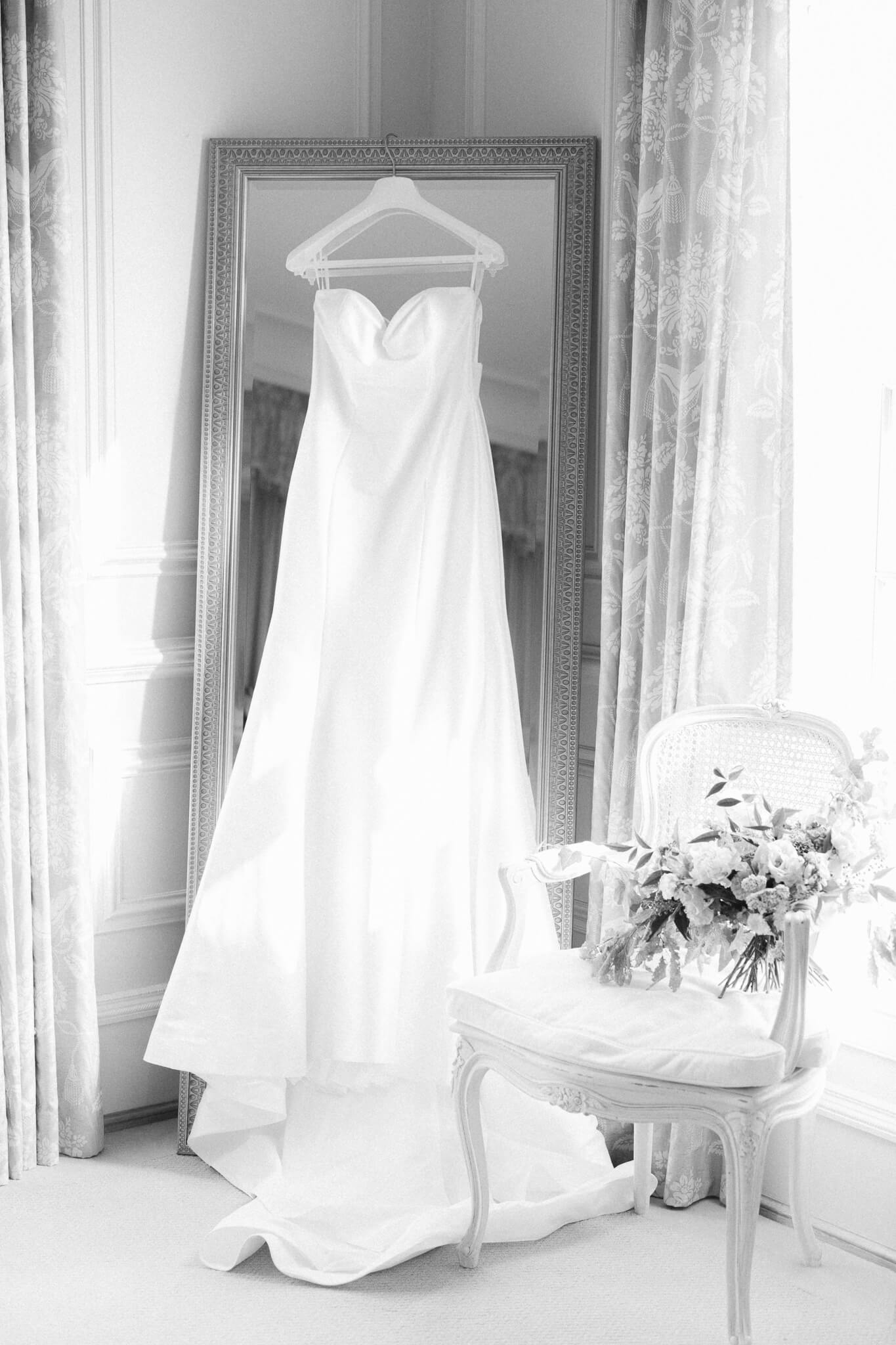 wedding gown hanging from mirror and bouquet on chair