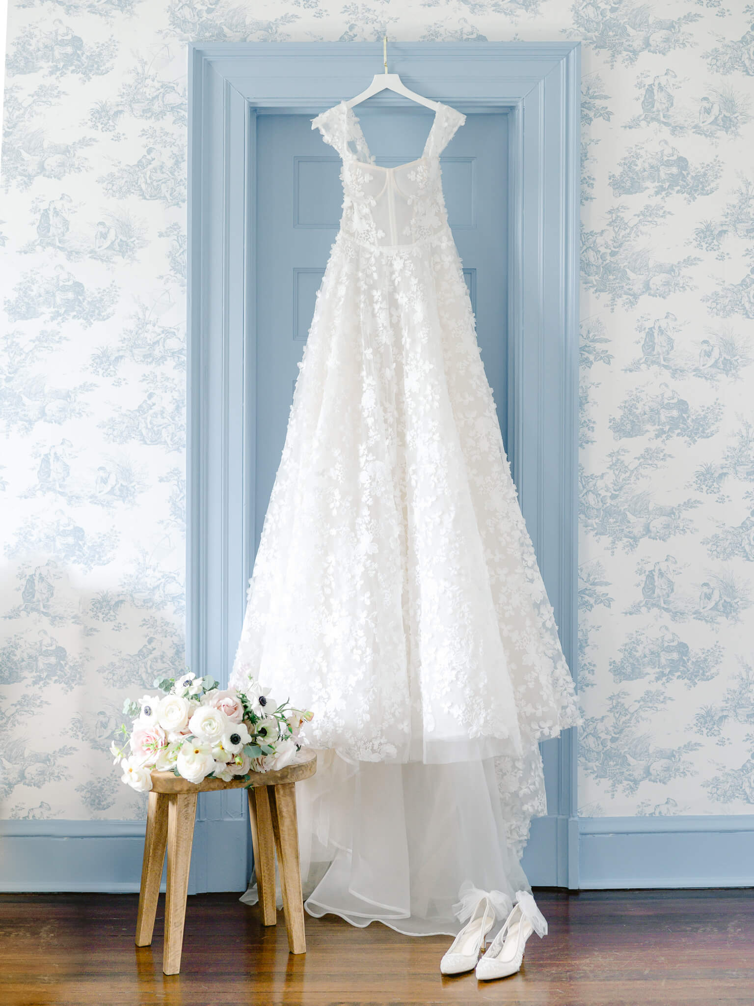 An Eisen Stein Bridal gown hanging on a blue doorframe in a blue toile wallpaper room with the bouquet on a wooden stool next to it.