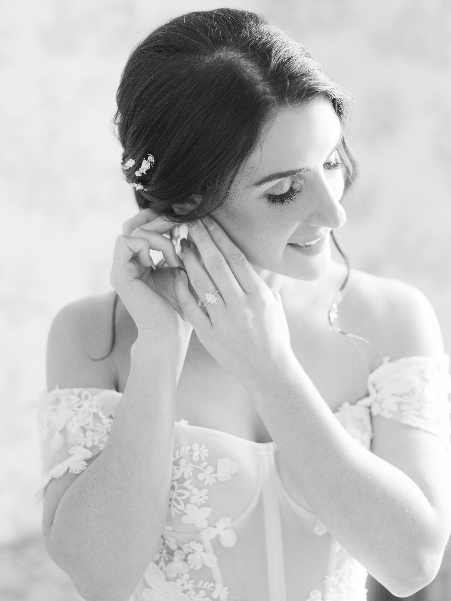 A black and white close up image of a bride in her wedding dress putting on her earrings.