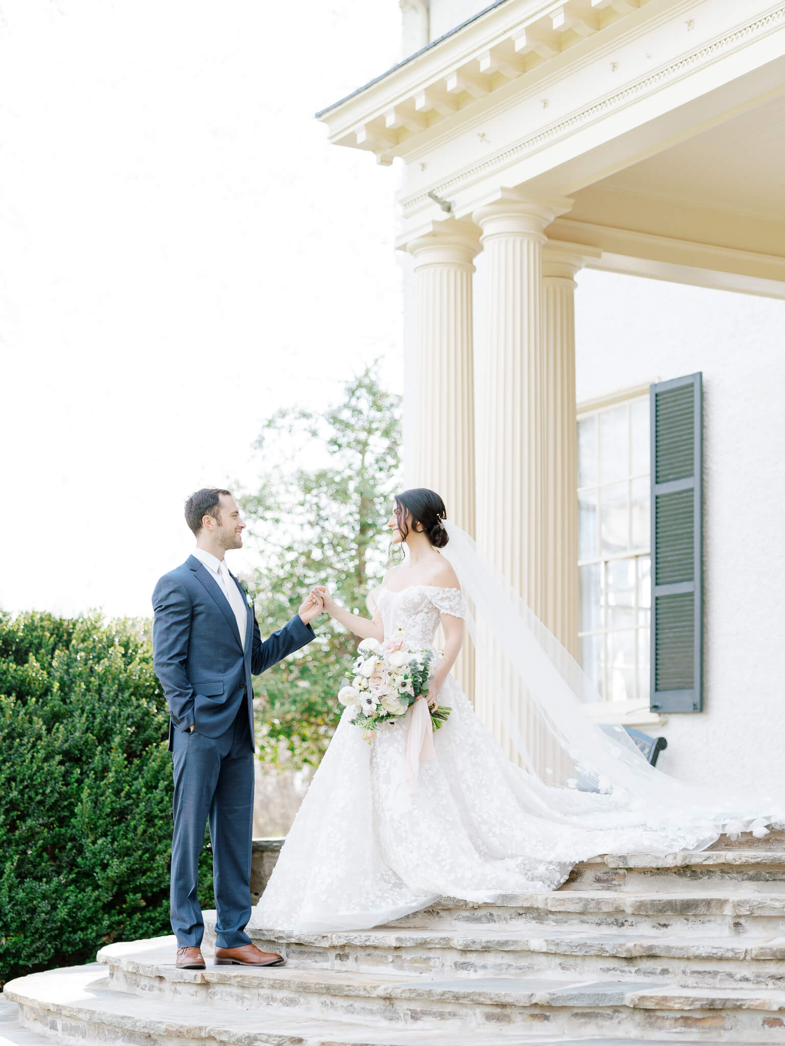 A groom in a blue suit leading a woman in an Eisen Stein Bridal gown down the back steps of Rust Manor House.