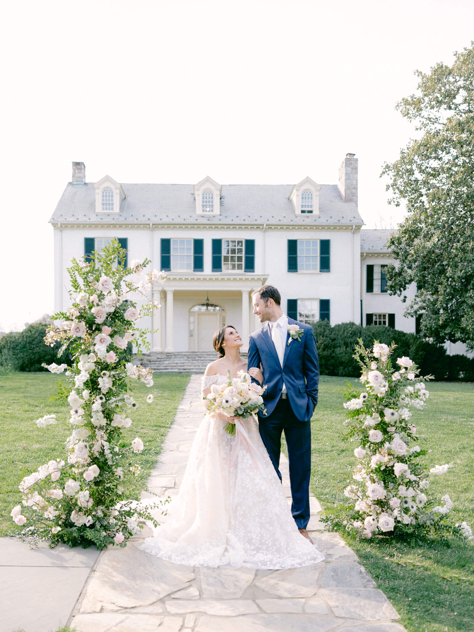 A newlywed couple in a suit and wedding dress standing between a flower arch in front of Rust Manor House.