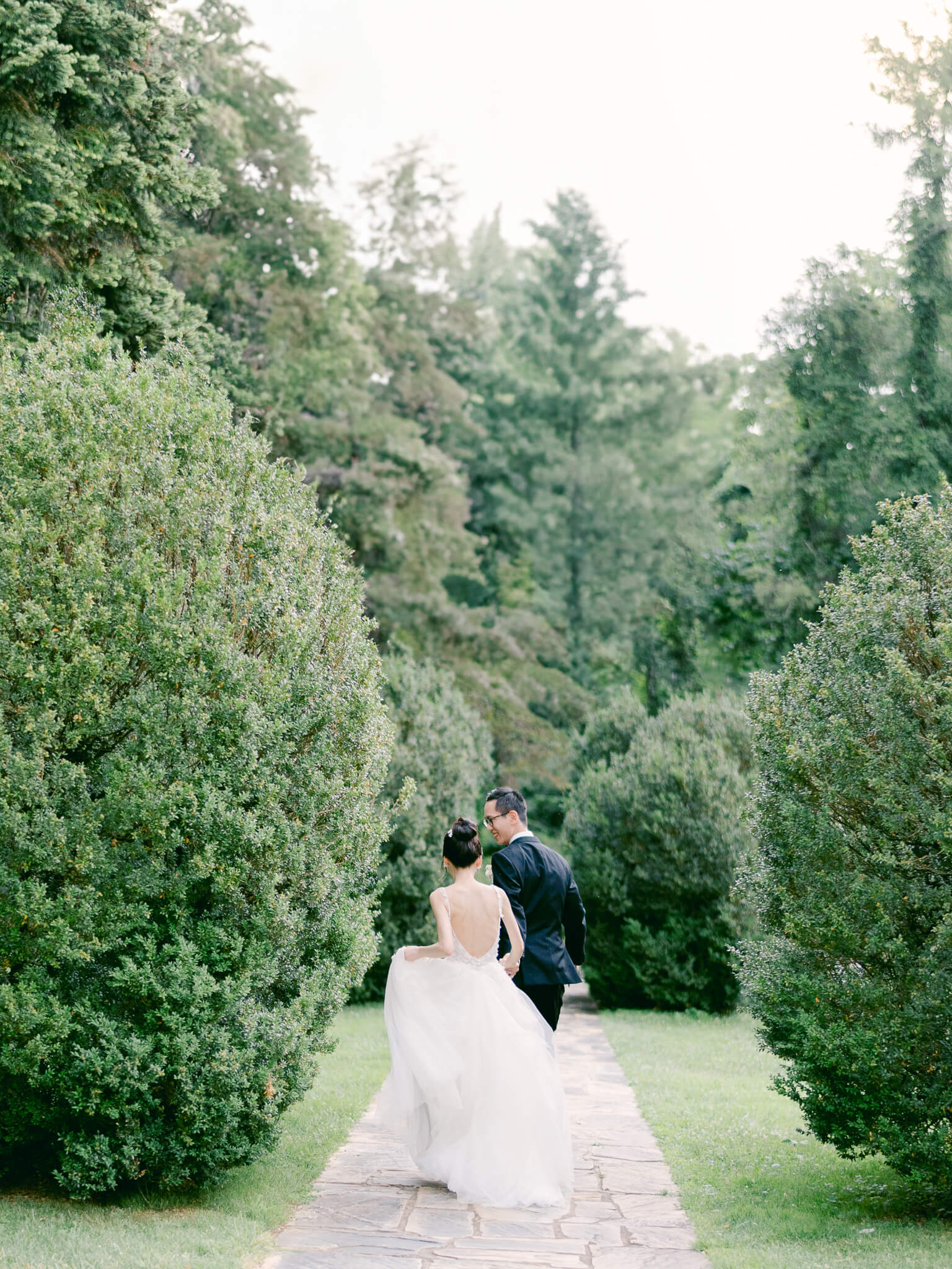 A bride and groom running away down a stone path between green shrubs and trees.