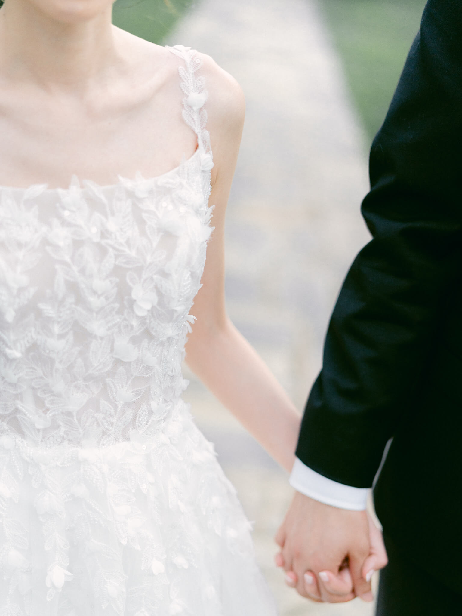 Closeup of a woman in a flower-detail wedding dress holding hands with her groom who is wearing a black suit.