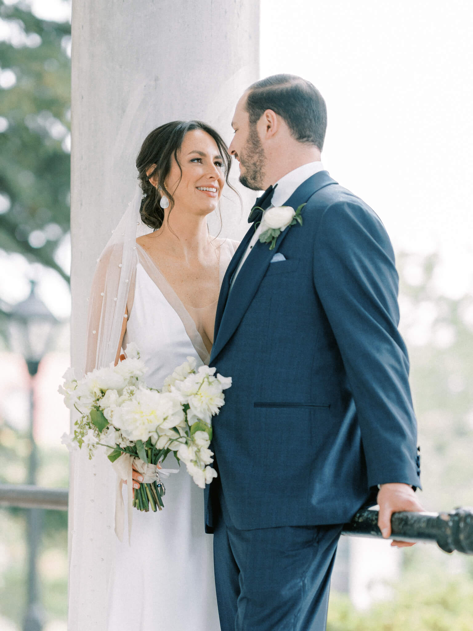 Bride holding her bouquet and groom in a blue suit close up together leaning against a railing and column