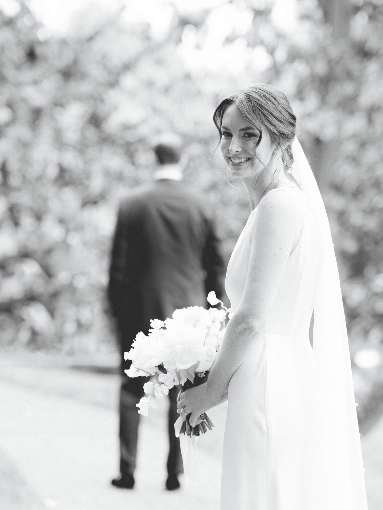 Black and white image of bride holding a bouquet and looking back with groom in the background