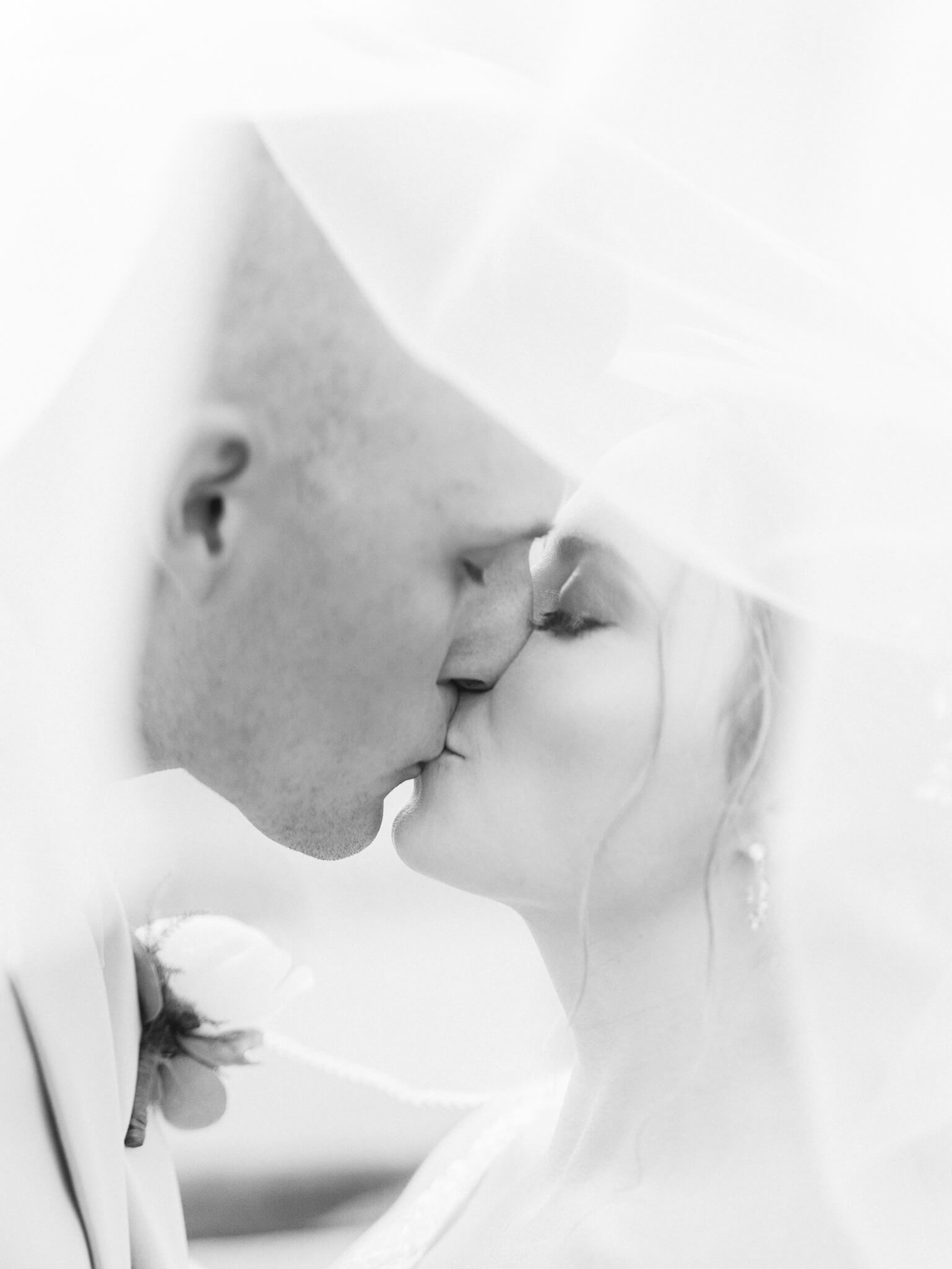 Close up of a bride and groom kissing under a veil in black and white