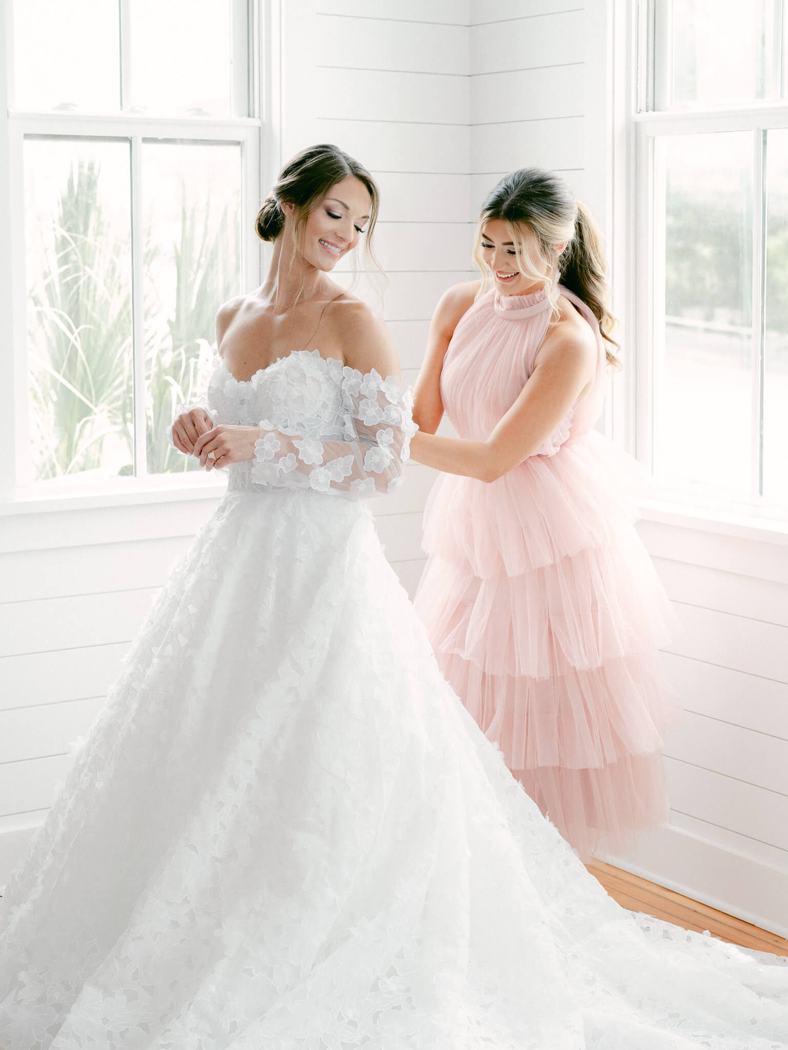 A bridesmaid in a blush tulle gown helping a bride into her off the shoulder wedding gown with sleeves.
