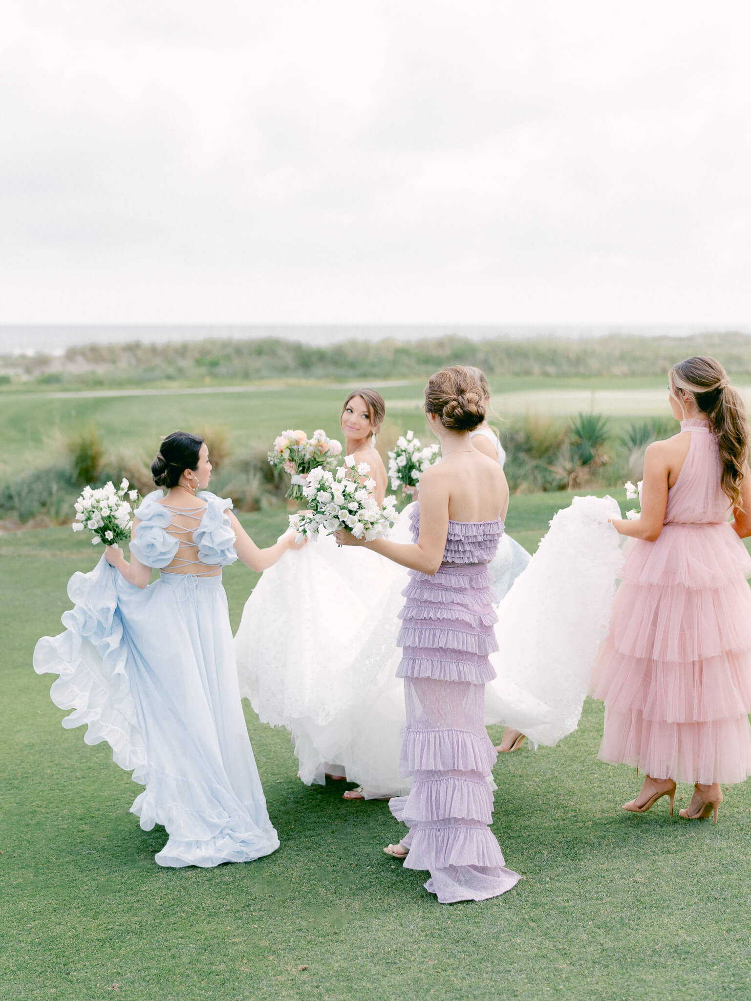 Three bridesmaids dressed in mismatched pastel blue, lilac and blush dresses walking across the golf course and helping the bride carry her gown.