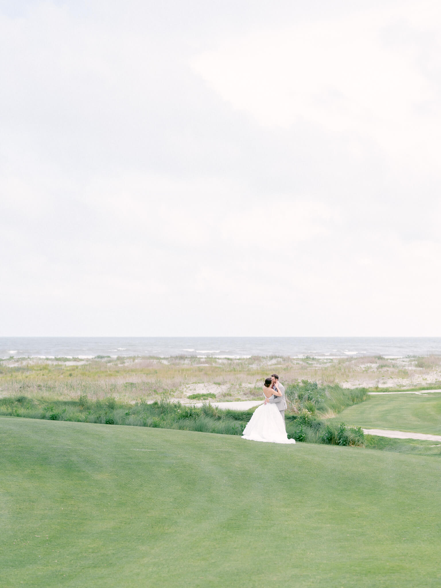 A bride and groom kissing in the distance on the Kiawah Island Ocean Course with the Atlantic Ocean and dunes in the background.