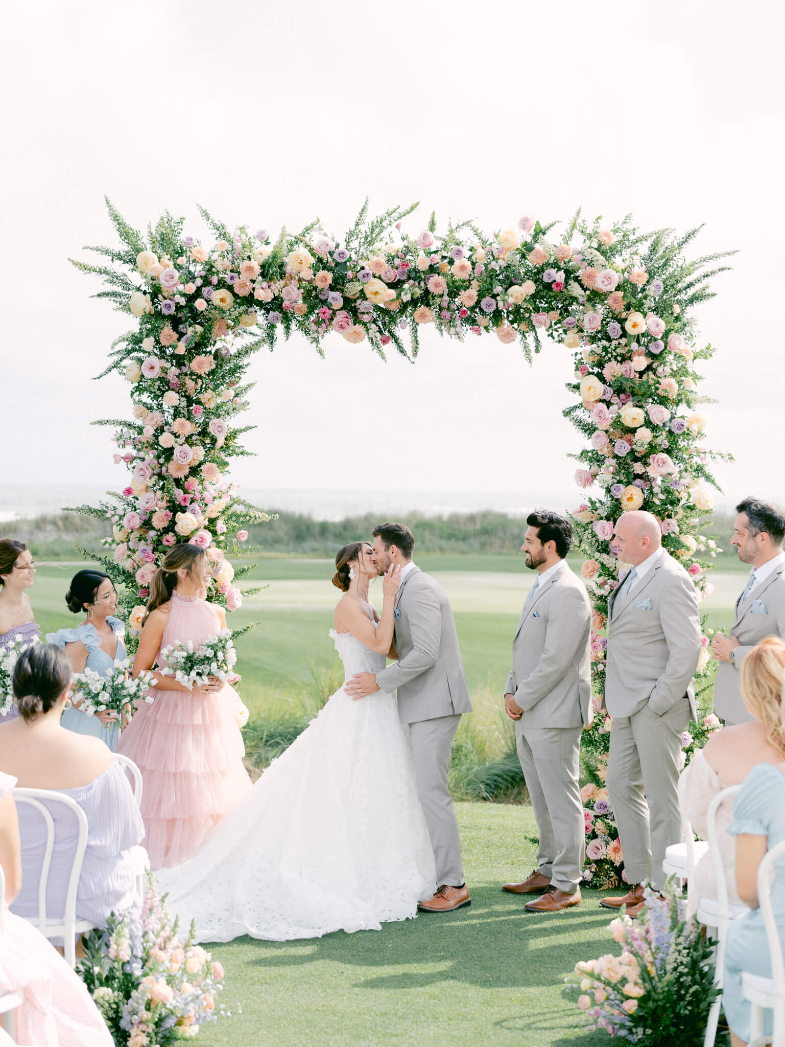 A bride and groom kissing for the first time in front of their guests under a large square floral arch.