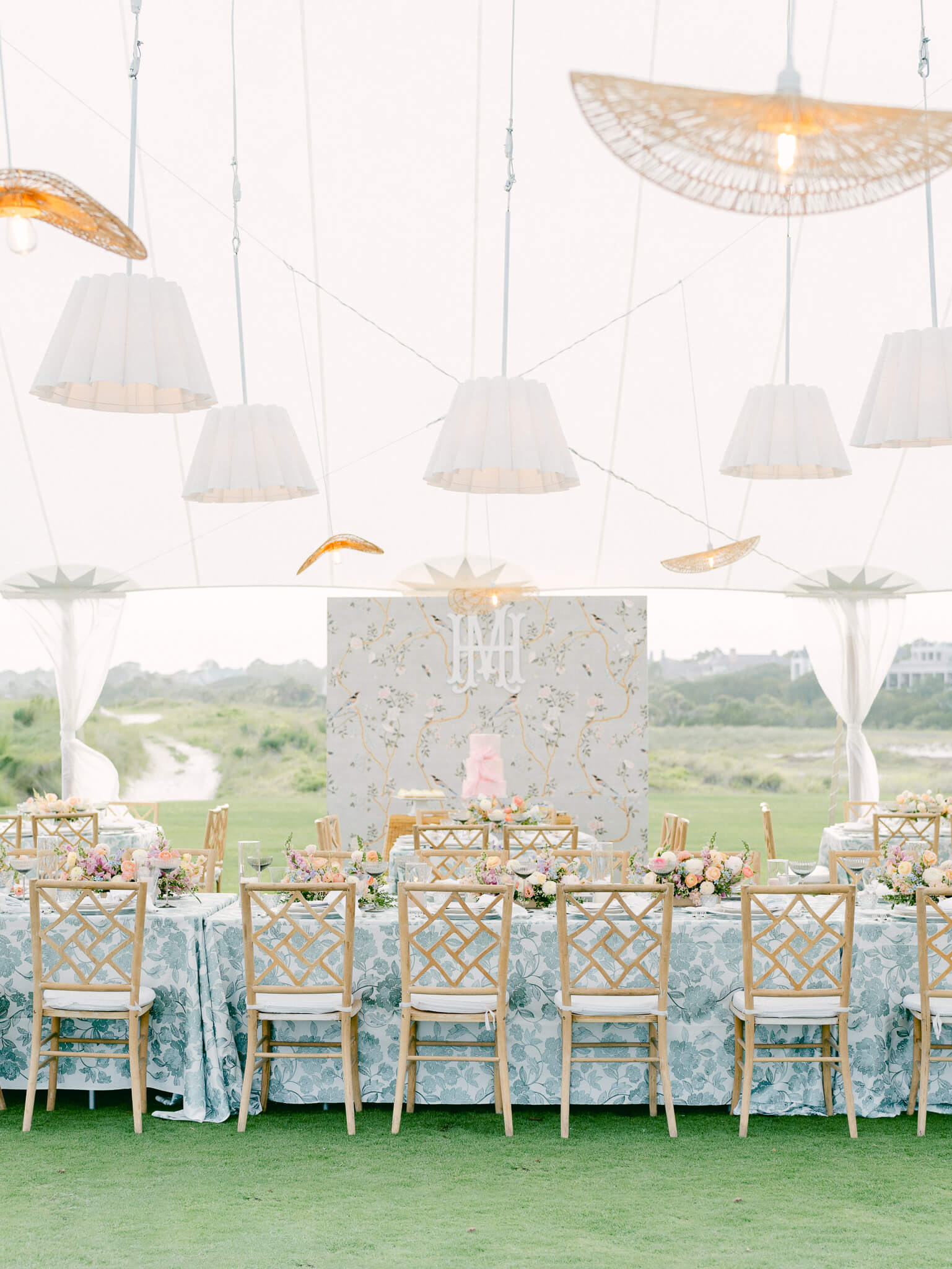 A long reception table with a blue tablecloth and chairs under a sail cloth tent with lamps hanging from above.