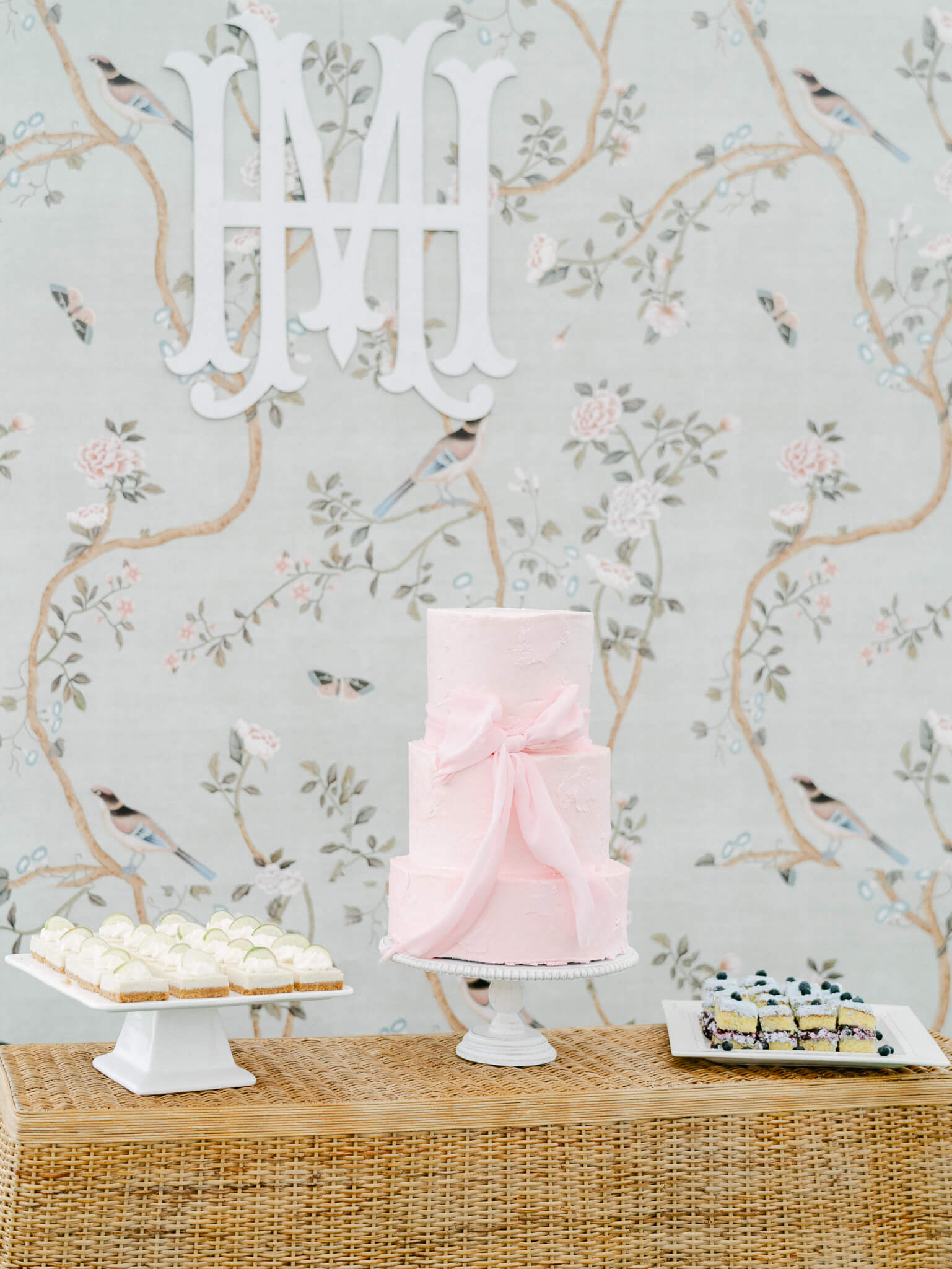 A pink three tier cake with a bow on it and desserts to each side in front of a mint chinoiserie monogrammed background.
