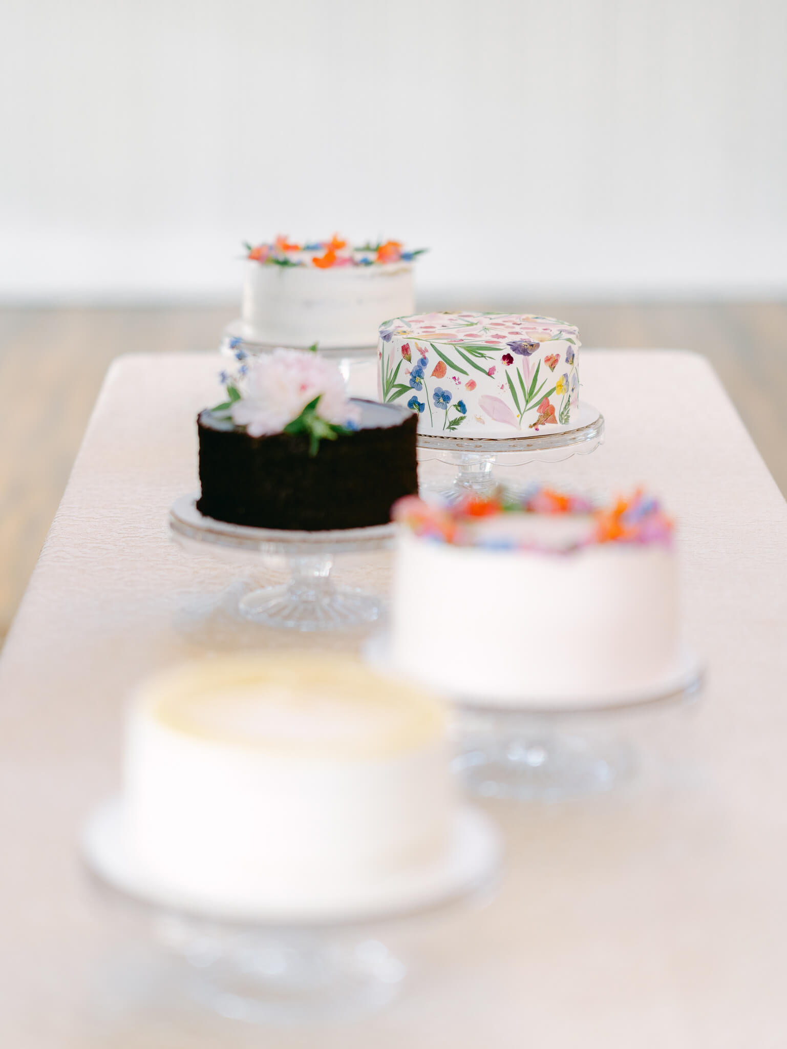 Five colorful round wedding cakes displayed on a blush tablecloth on a long table.