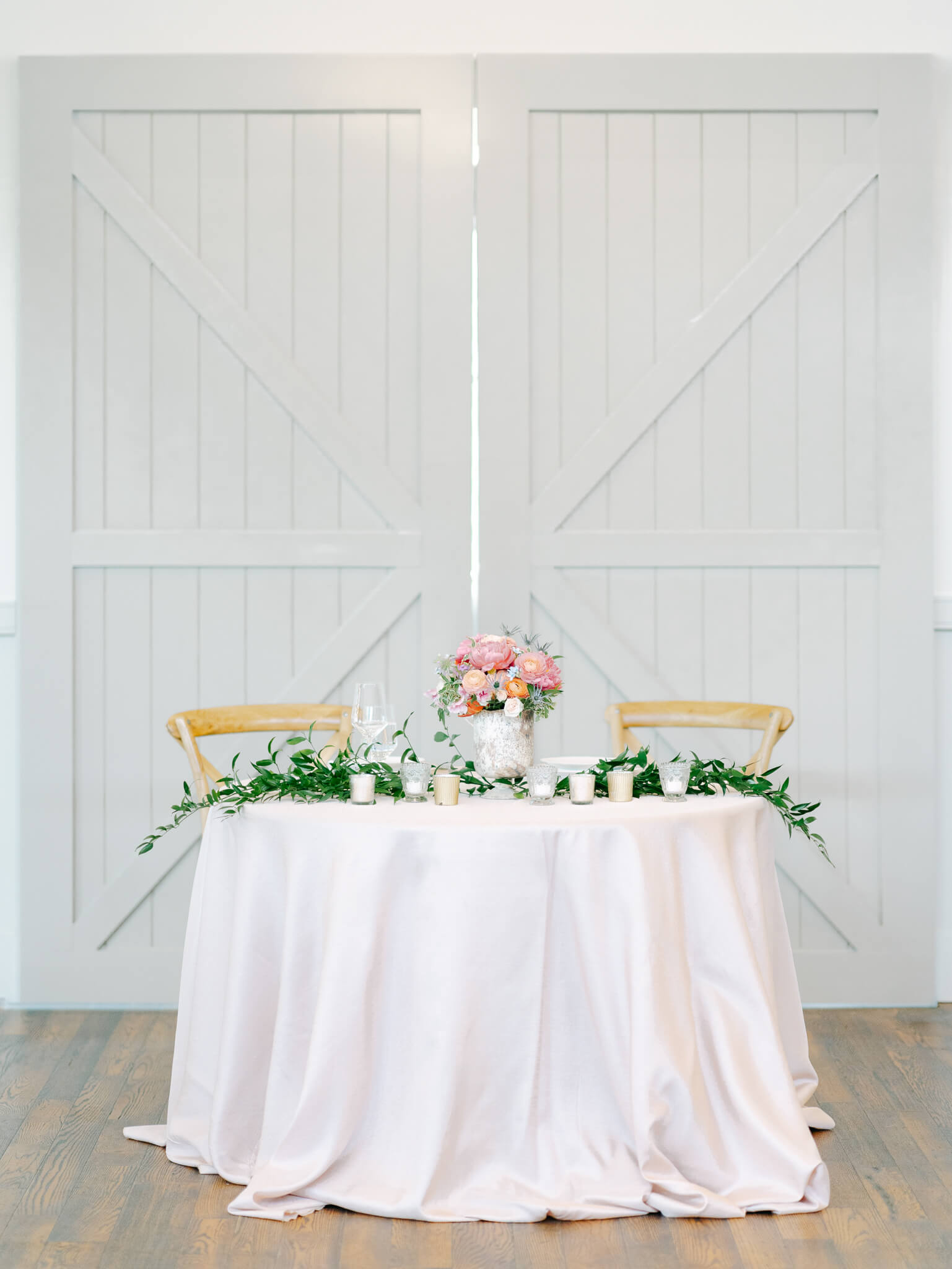 A sweetheart table decorated with a blush table cloth, colorful bouquet and greenery in front of grey barn doors inside a reception space.