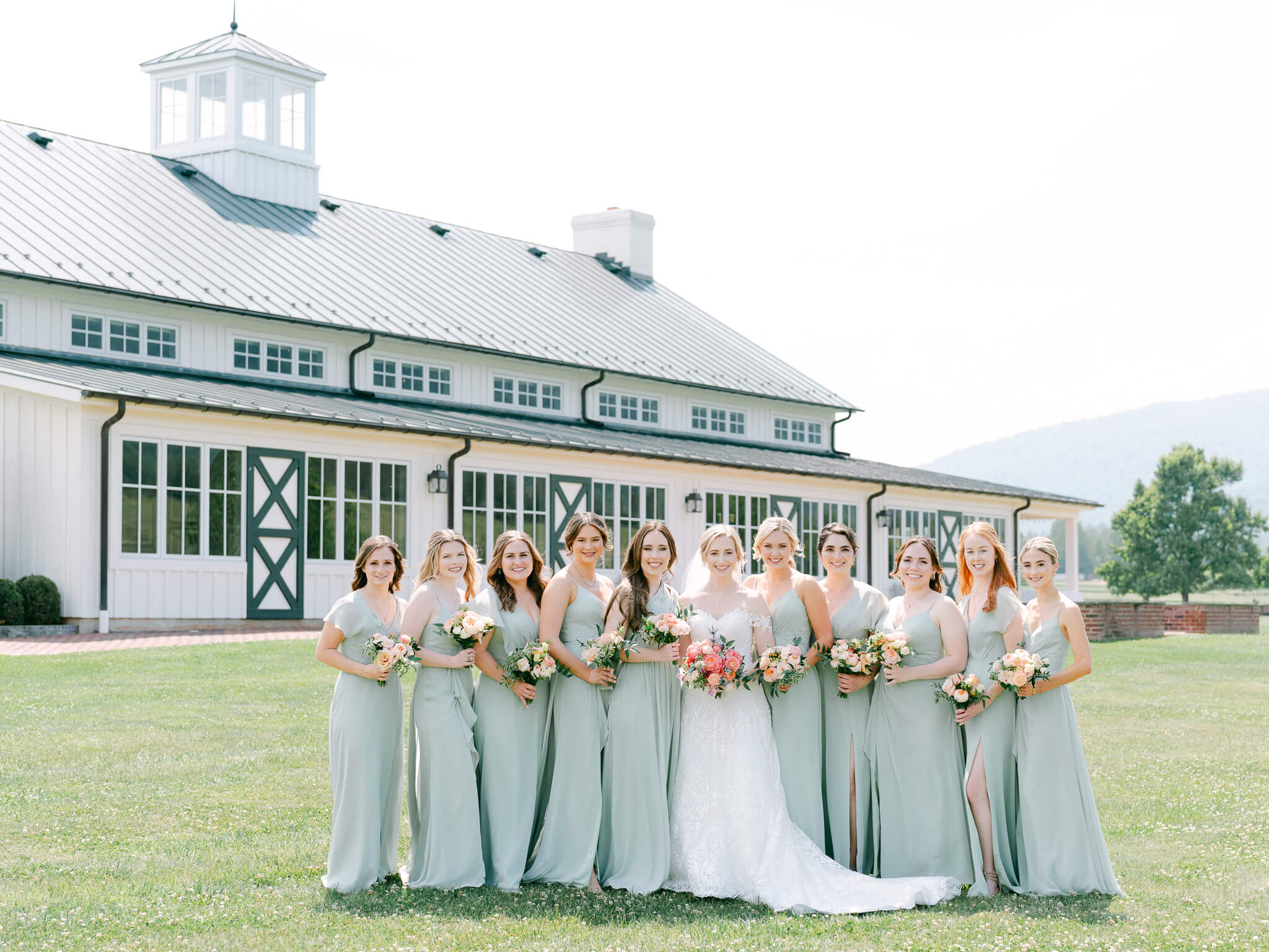 A bride and her bridesmaids in sage dresses lined up on the lawn in front of King Family Vineyard's white and black building.