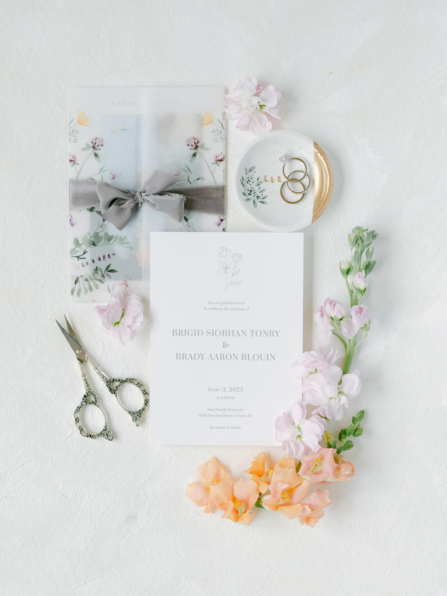 A wedding flatlay on a white mat with an invitation, rings in a small dish, colorful flowers and small scissors.