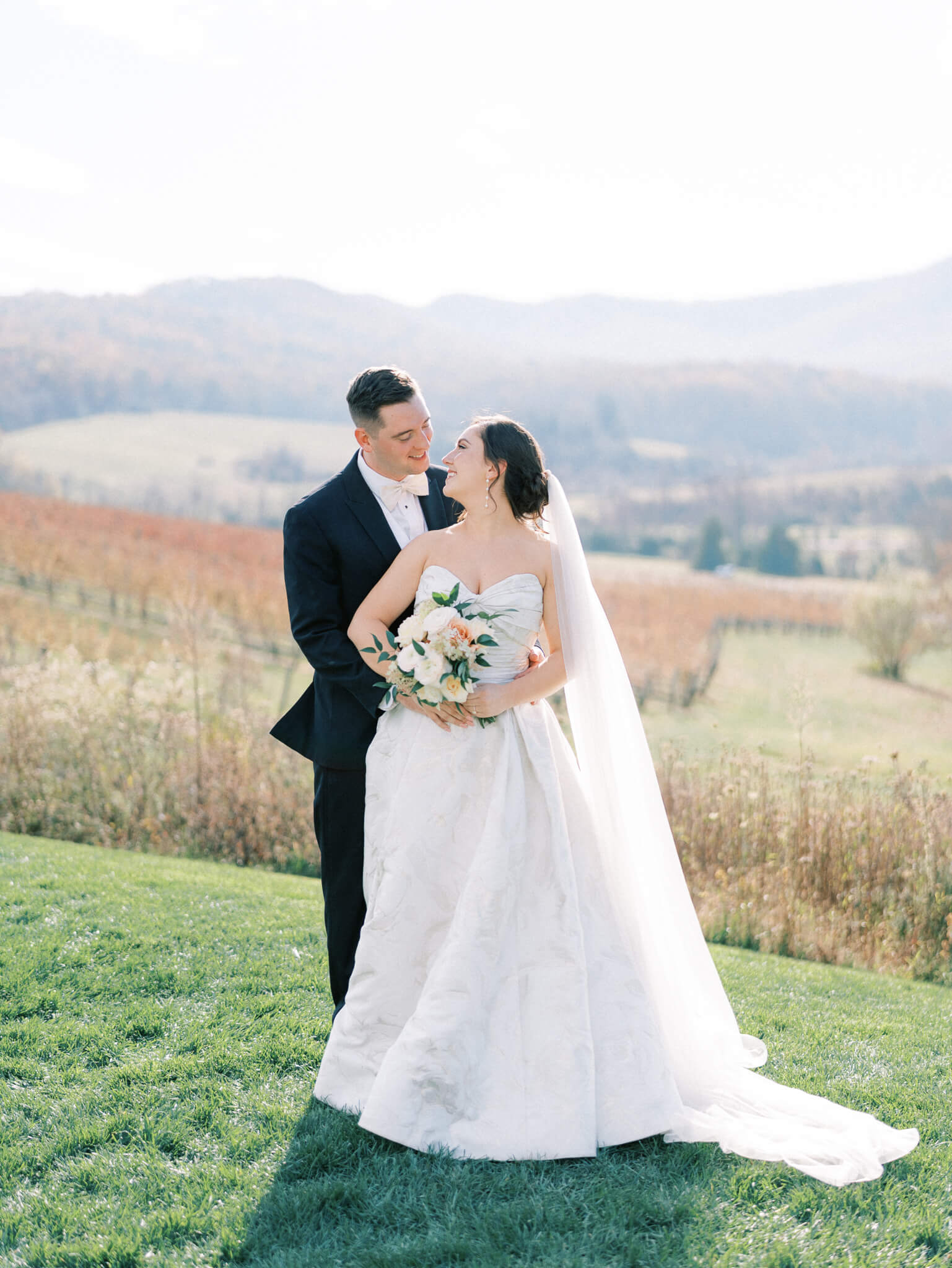 A groom standing behind a bride and embracing her while she leans in to kiss him in front of the picturesque mountain views at a Pippin Hill wedding.