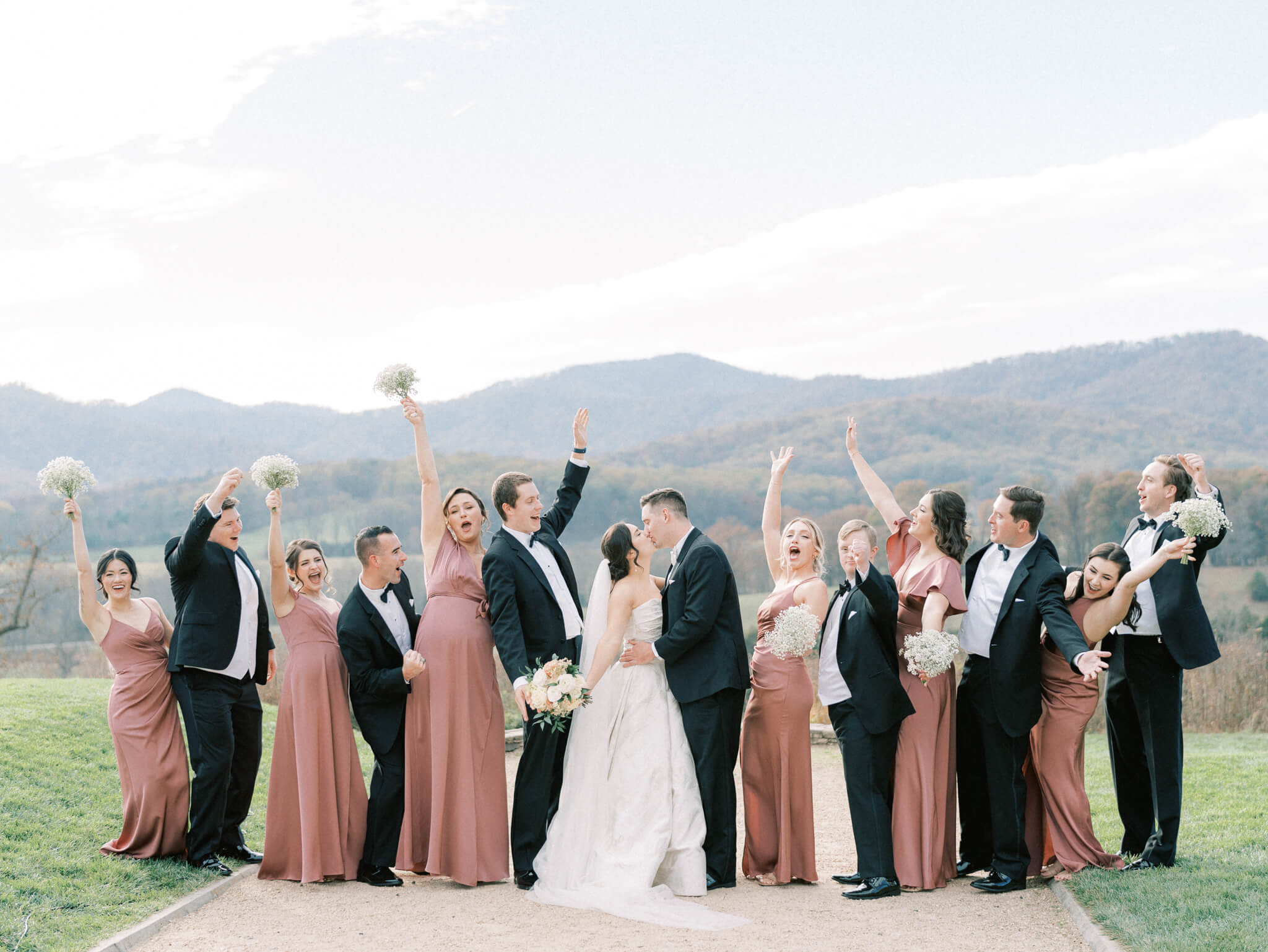 An intermingled wedding party, wearing mauve dresses and black suits, cheering for the bride and groom who are kissing in the middle.
