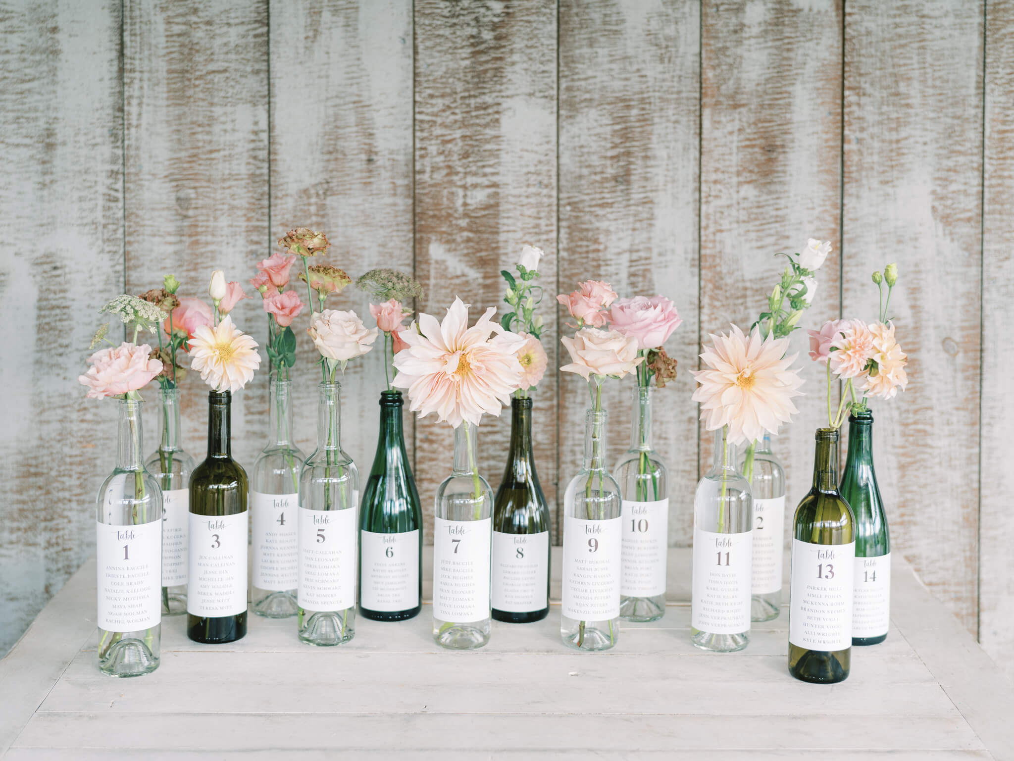 A seating display made of green, brown and clear wine bottles with white labels and peach blush dahlias and roses at Pippin Hill Vineyards.