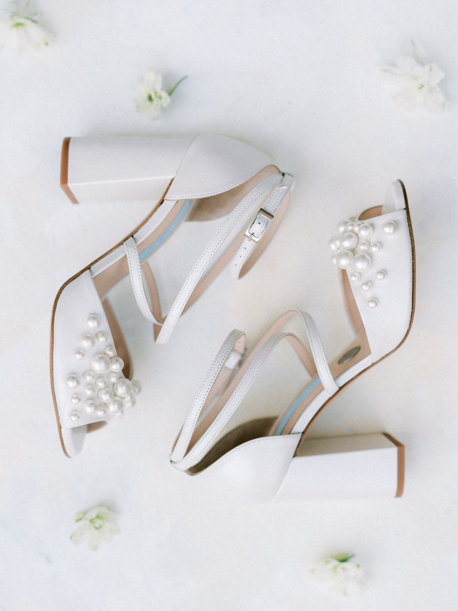 Flatlay of a pair of white bridal heels with pearls on a white background with white flowers.