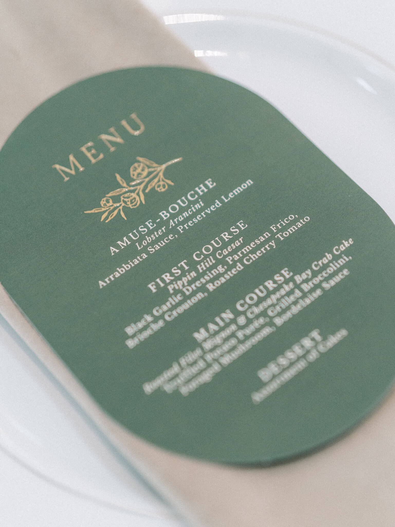 Close up of a green oval wedding dinner menu with gold and white writing on top of a tan napkin and white plate.
