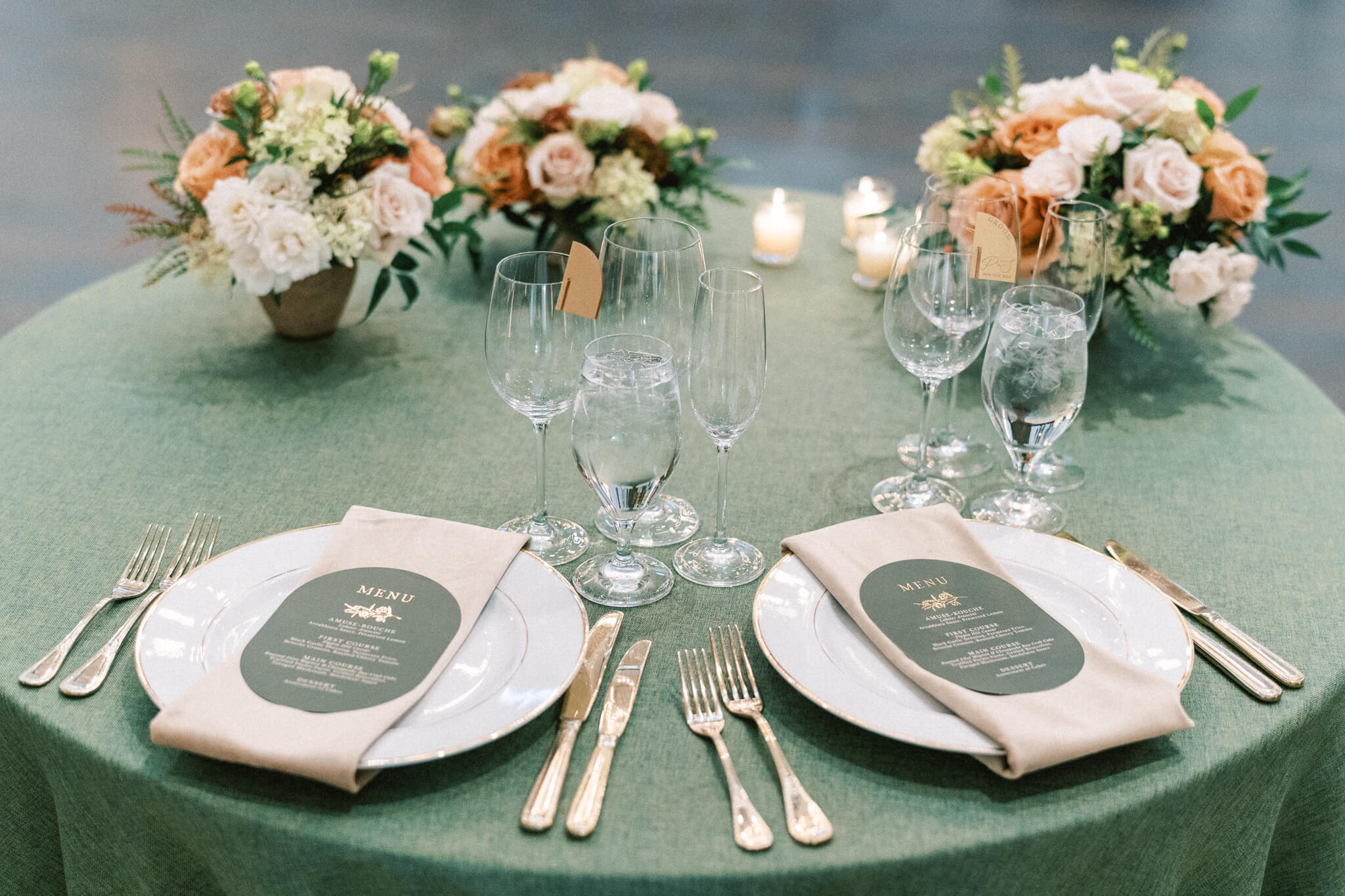 Closeup of a sweetheart tablescape with plates, menus, flatware, glasses, candles and florals on a green tablecloth.