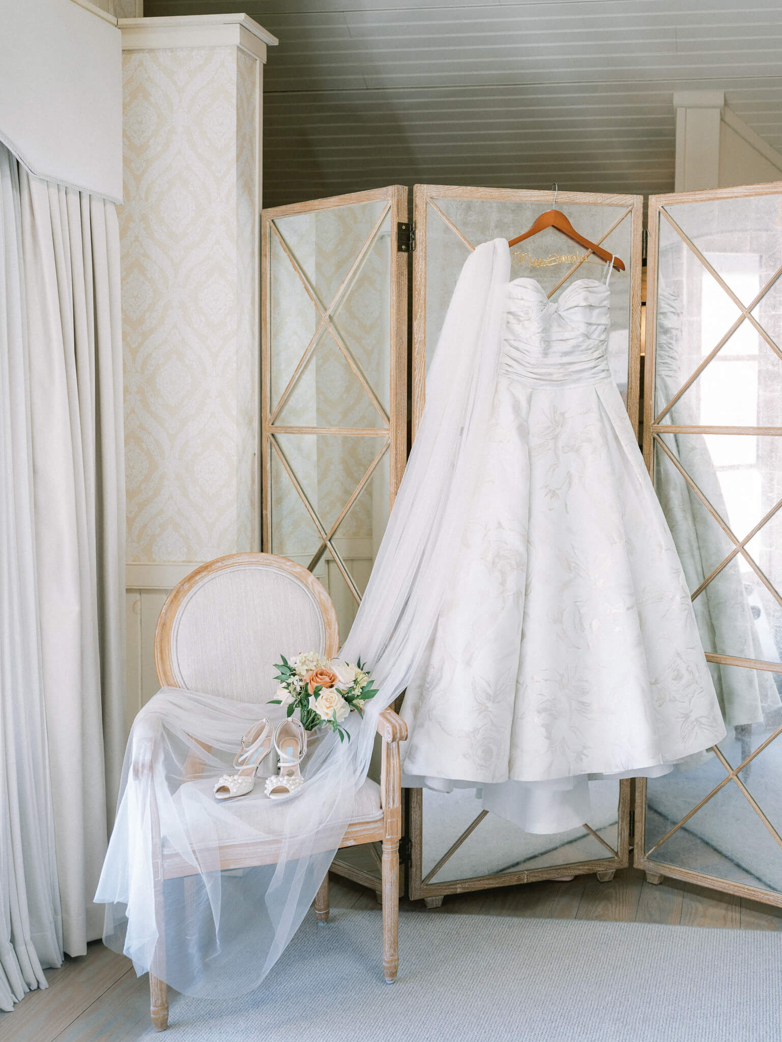 A floral wedding gown hanging from a mirrored divider next to a chair with the bride's veil, bouquet and shoes.