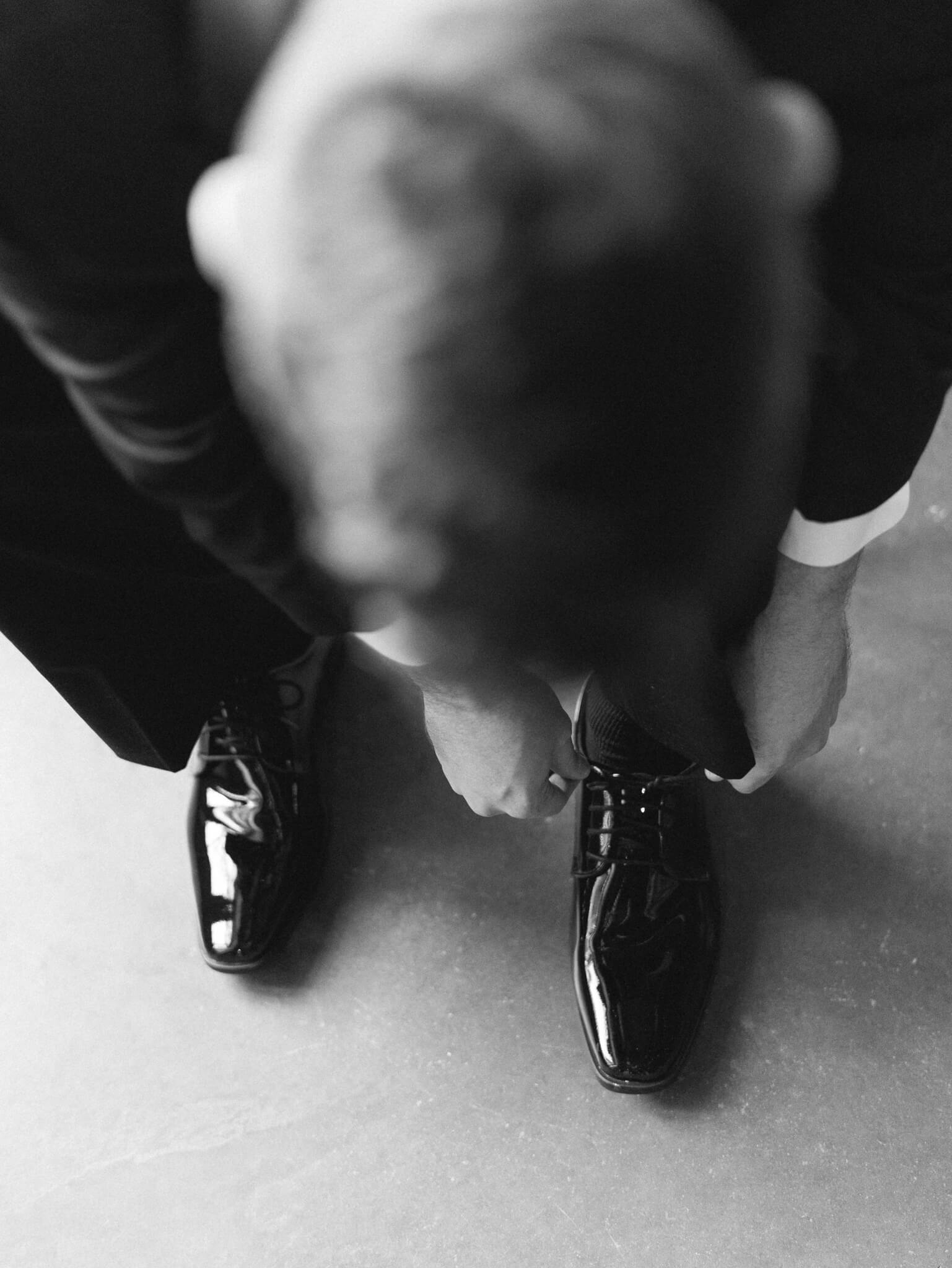 Black and white image from above of a groom in a tux tying his shoe.