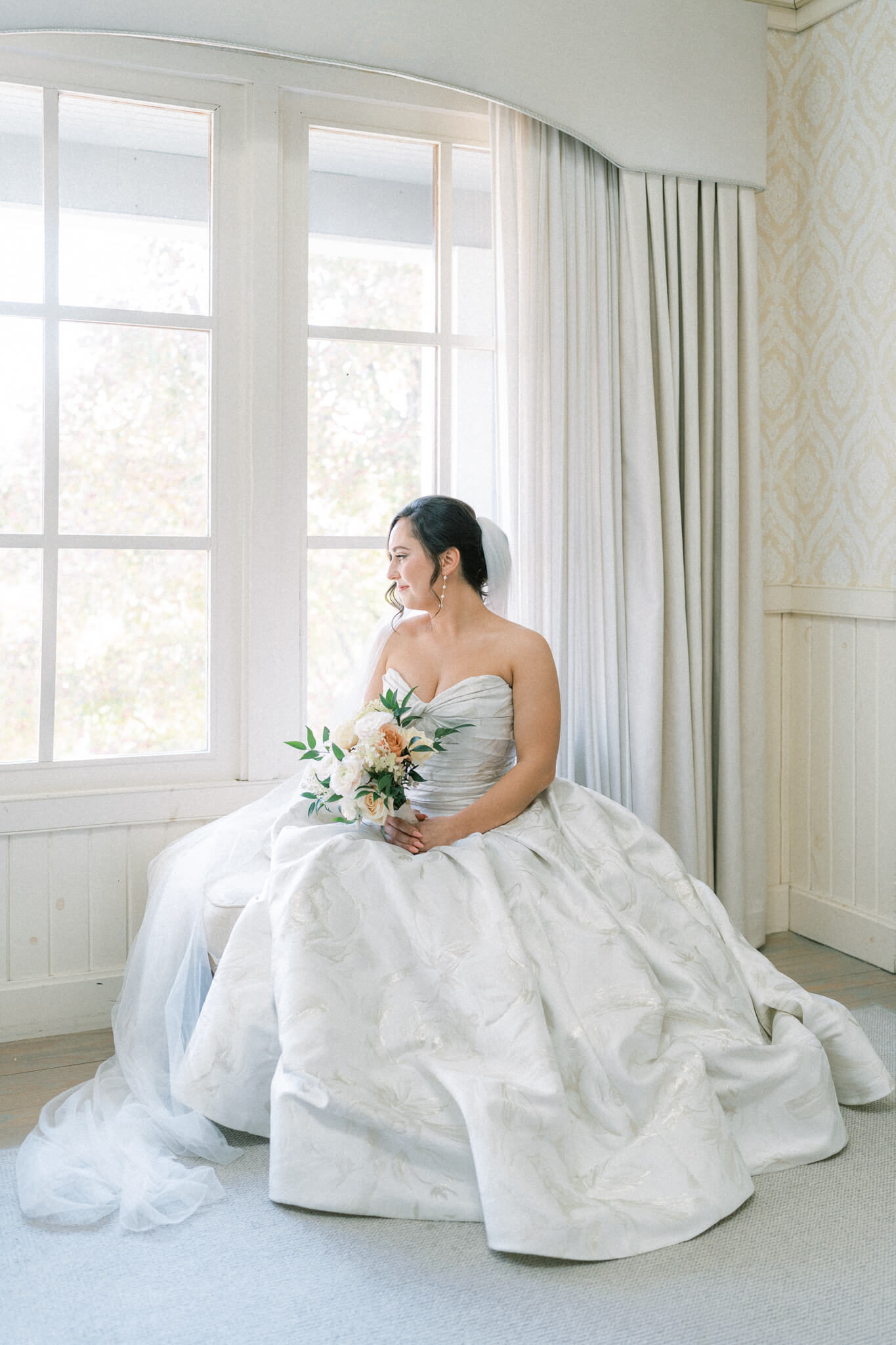A bride in her wedding gown sitting on a chair in front of a window looking out.