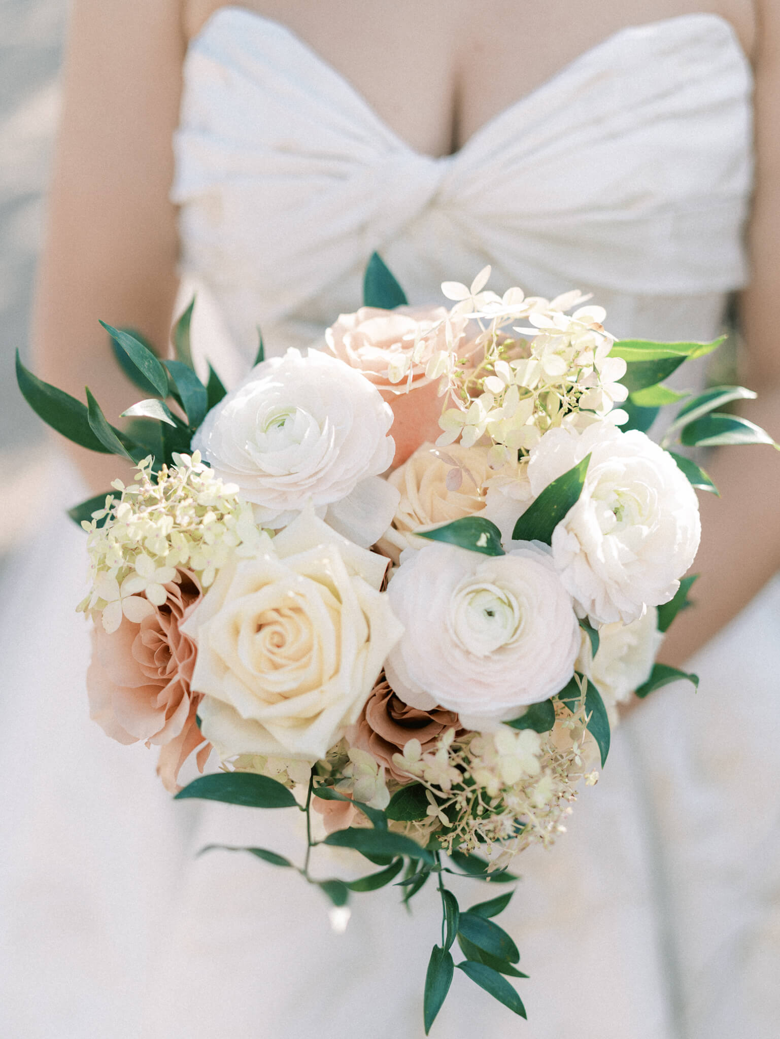 Closeup of a bride holding a bouquet of cream and peach colored roses with white ranunculus and hydreangeas.