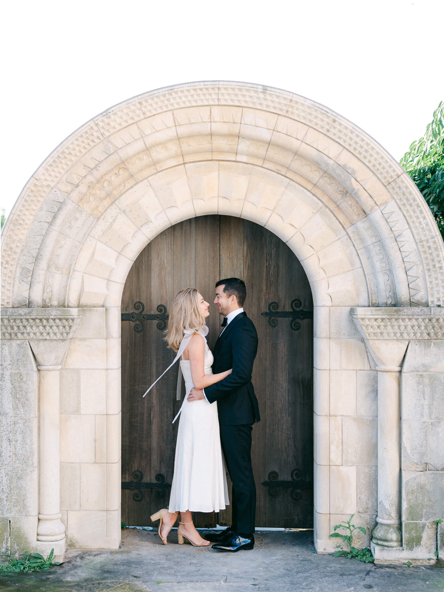 An engaged couple embracing in front of an arched door at Bishop's Garden.