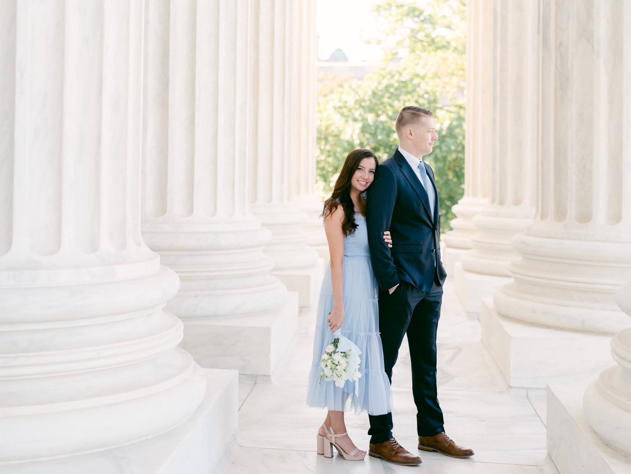 A woman in a blue dress holding a bouquet of white flowers leaning against her fiancé between the columns of the Supreme Court.