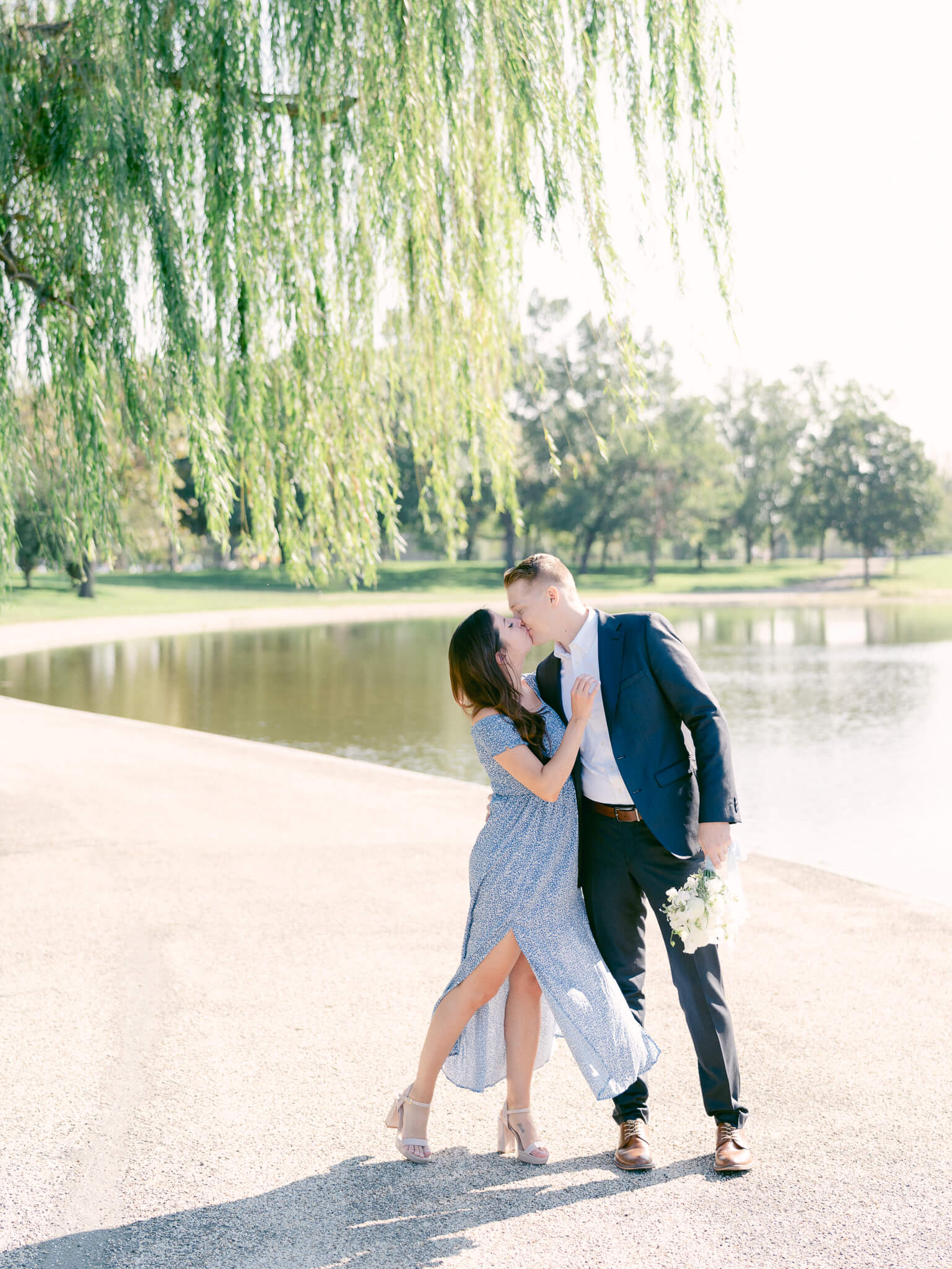 A man in a blue suit kissing a woman in a blue dress underneath a willow tree by a pond.