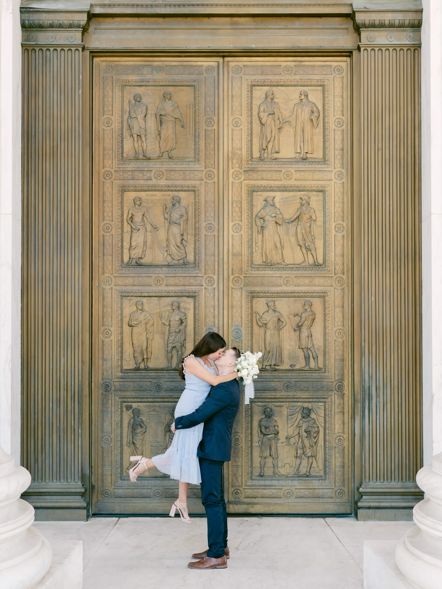 A man lifting his fiancée up and kissing her in front of the doors of the Supreme Court in Washington, D.C.