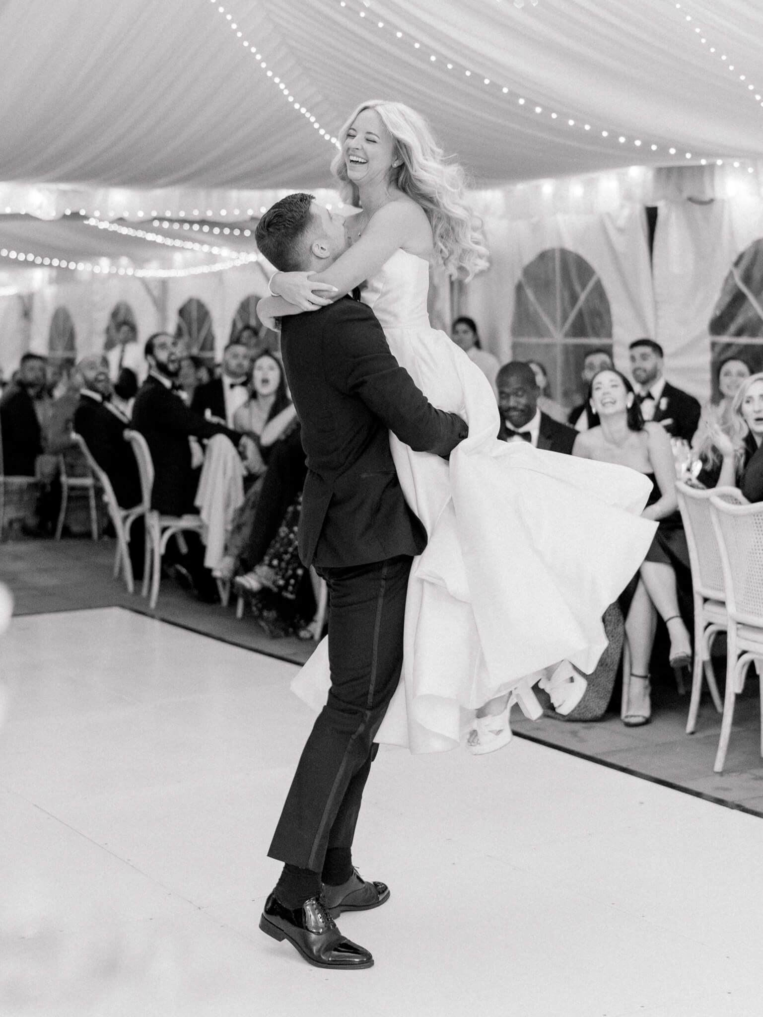 A groom picking up his bride and spinning her around while guests are watching their first dance in the tent at River Farm Wedding Venue.