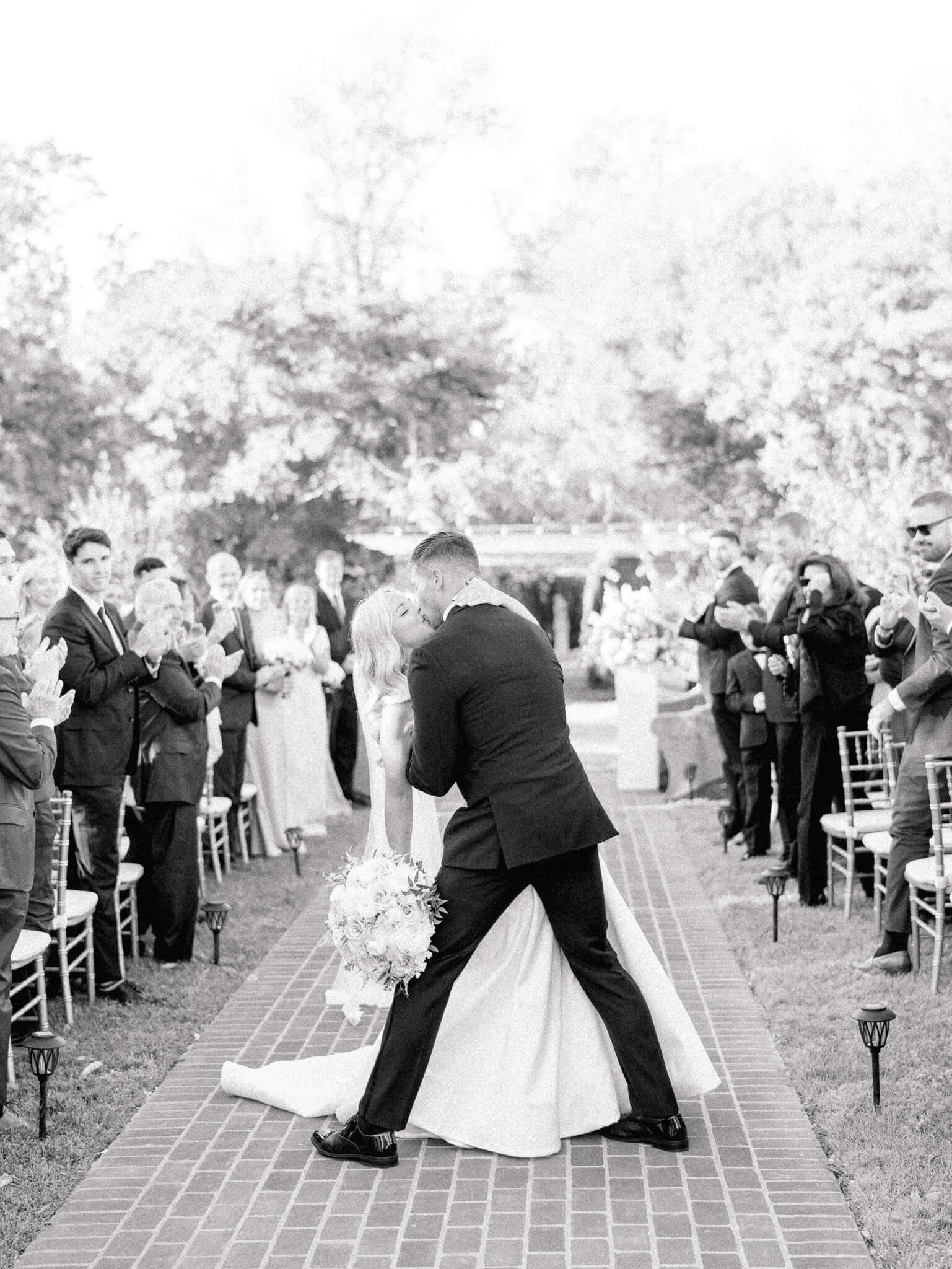 Black and white image of a groom kissing his bride and dipping her back at the end of the ceremony isle while guests watch and clap.