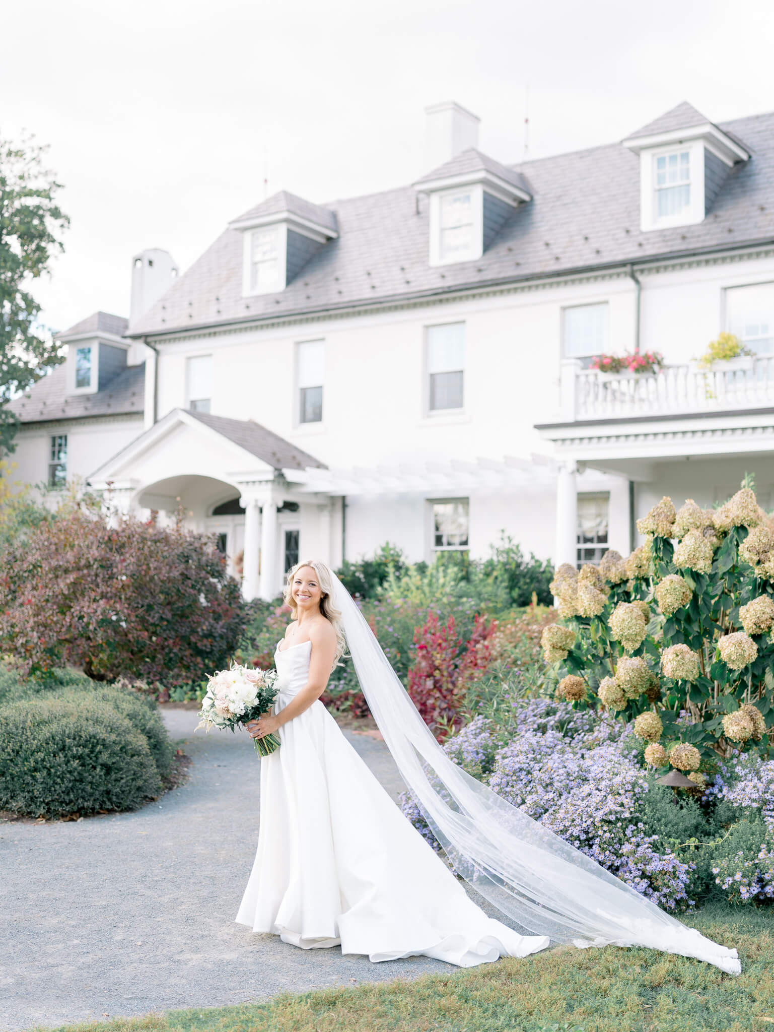 A bride in her wedding gown and cathedral length veil standing in front of River Farm Wedding venue holding her bouquet.