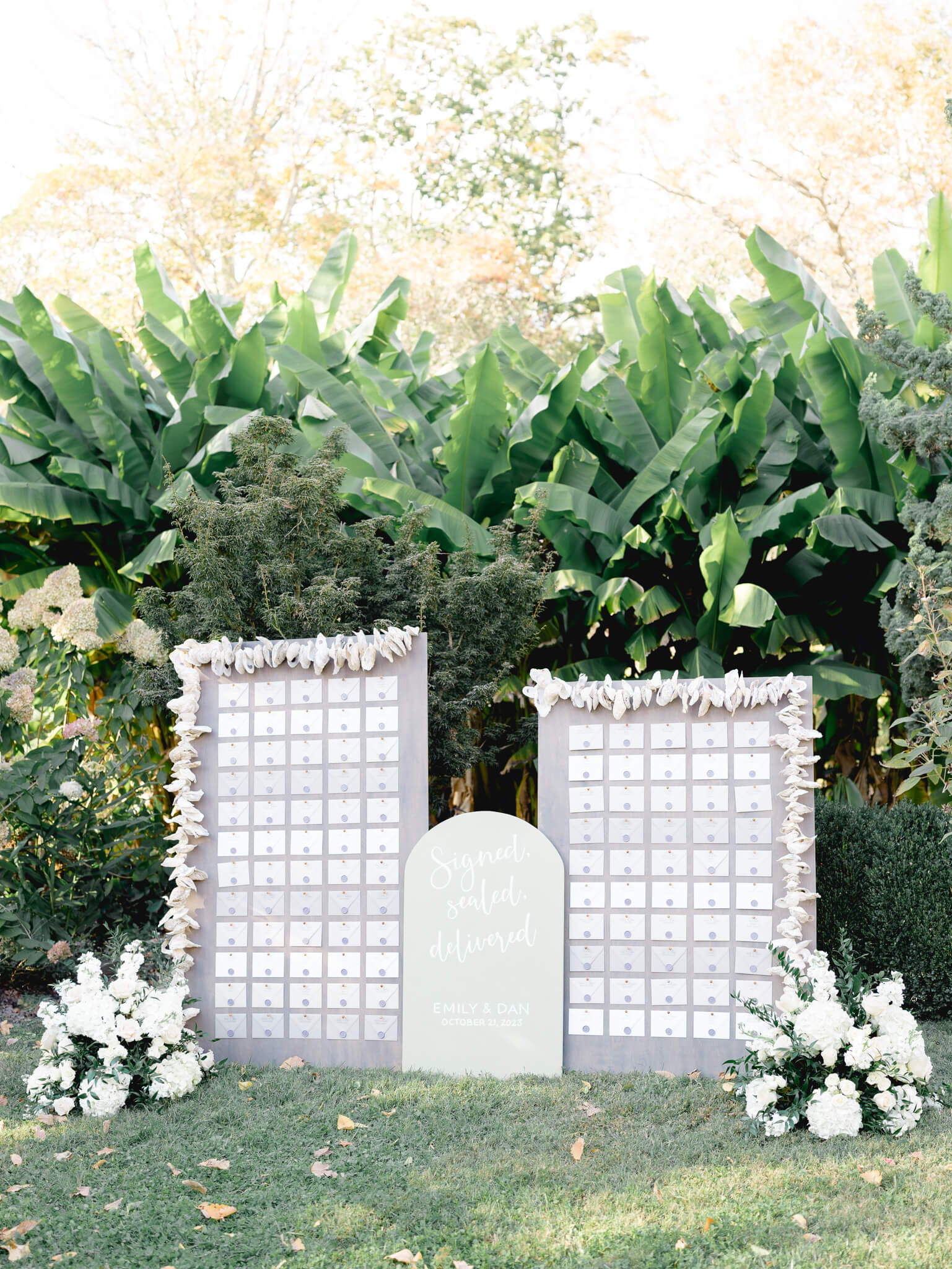 A gray escort chart made with oysters and envelopes standing in front of the banana plants at River Farm Wedding Venue.