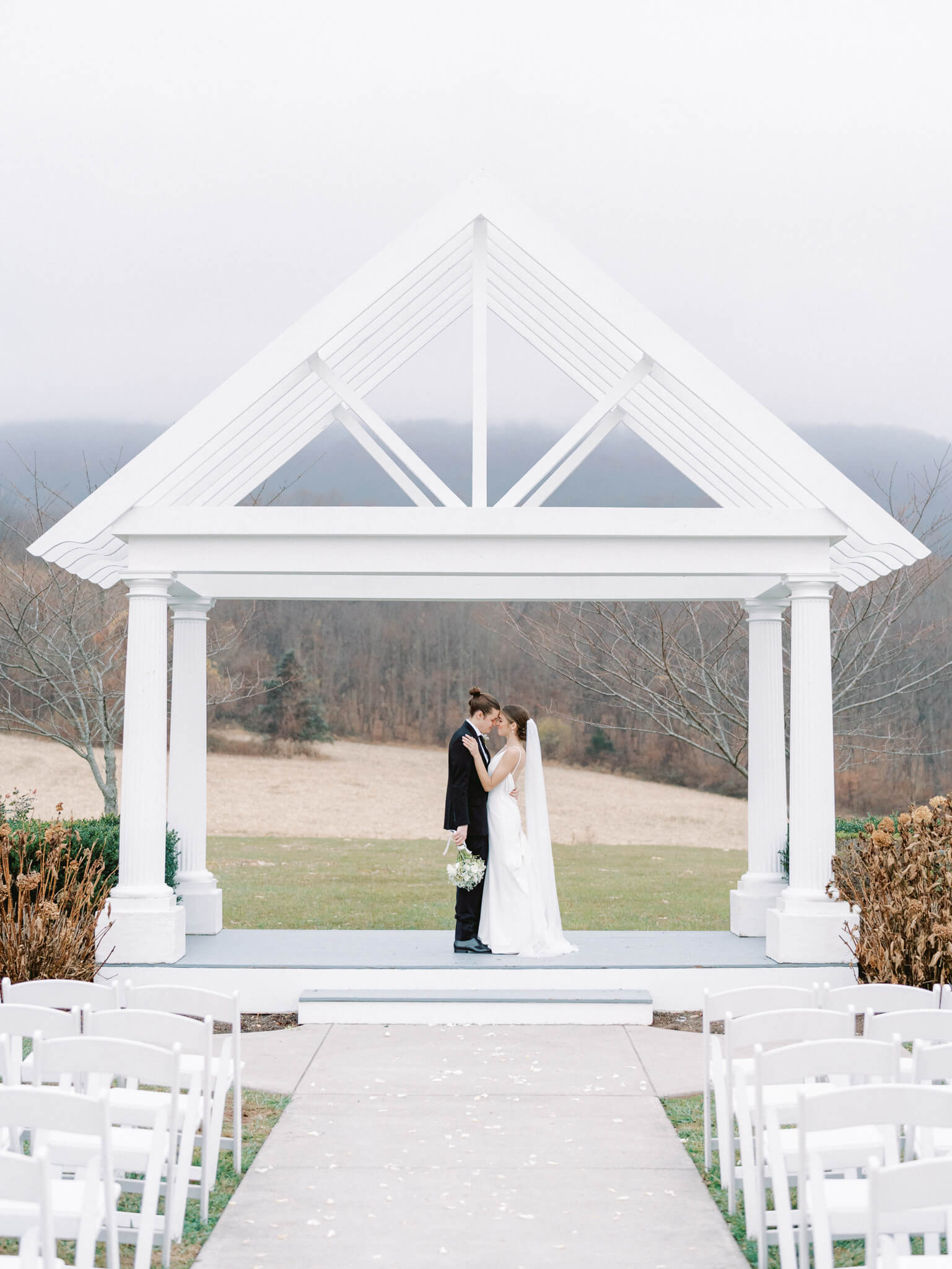 A bride and groom standing under their white arbor with their empty white ceremony chairs and fog in the background.
