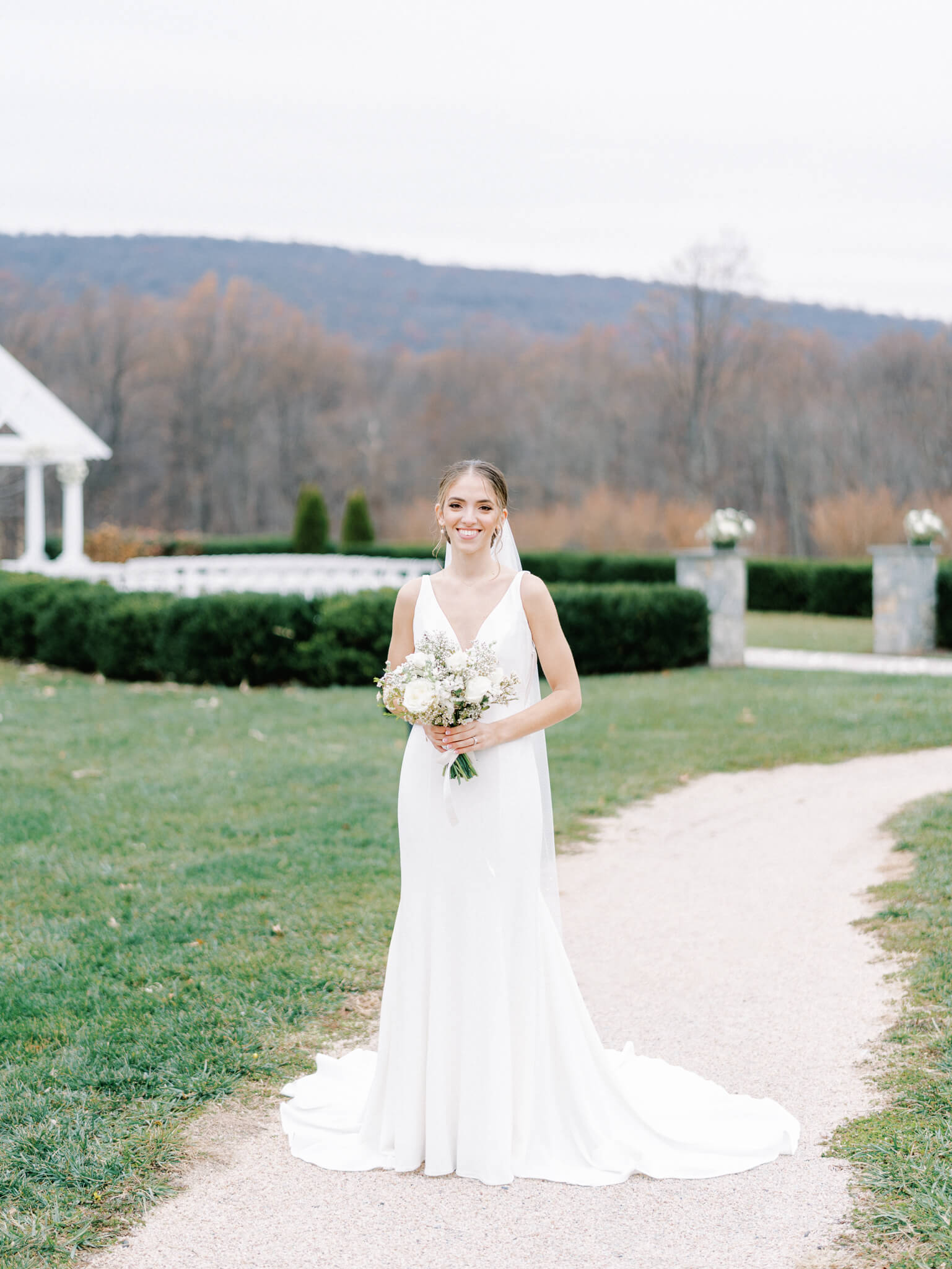 Full length portrait of a bride in her sleek gown holding her white bouquet in front of her ceremony site.