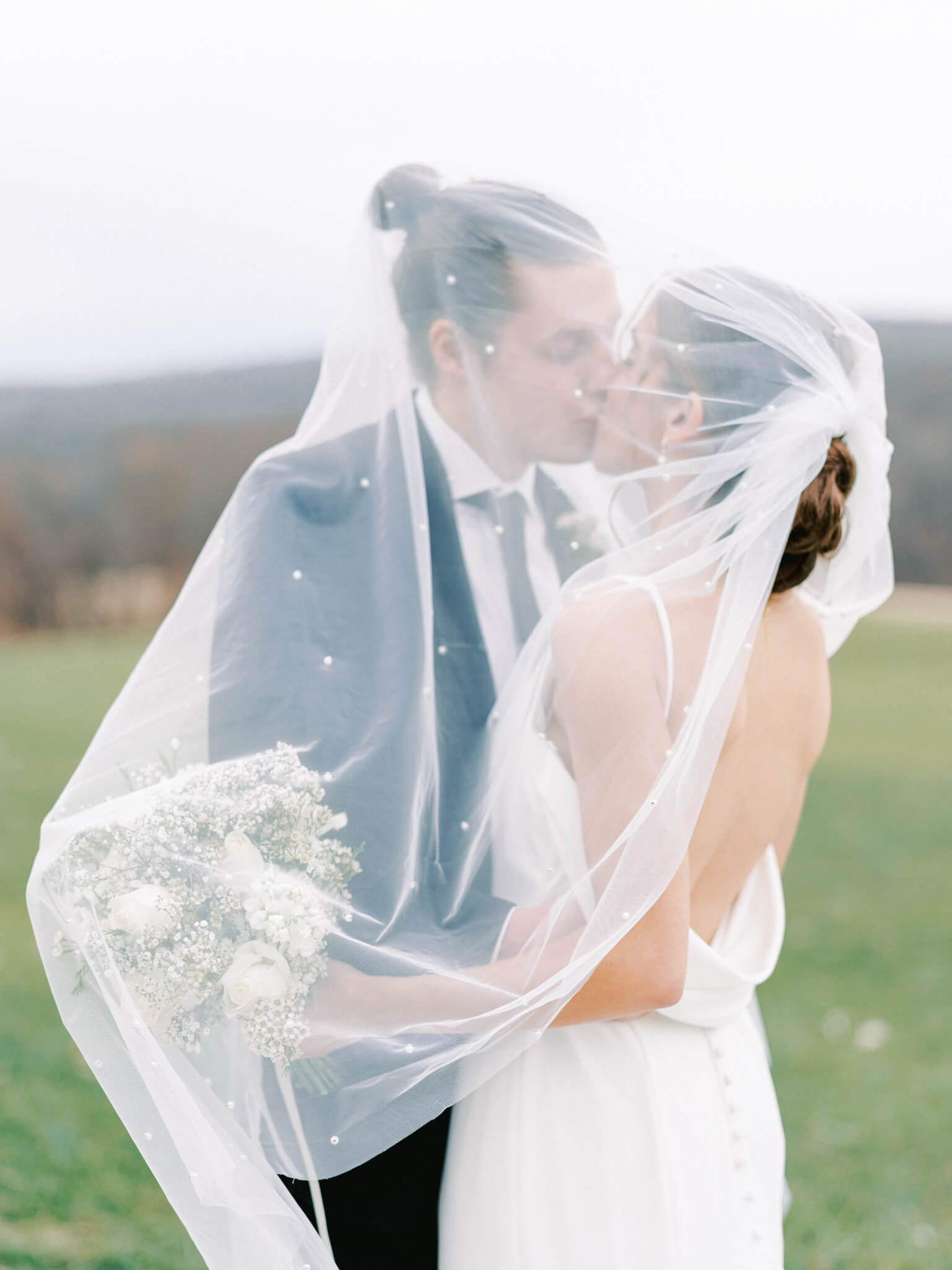A bride and groom kissing under a white veil with pearls.