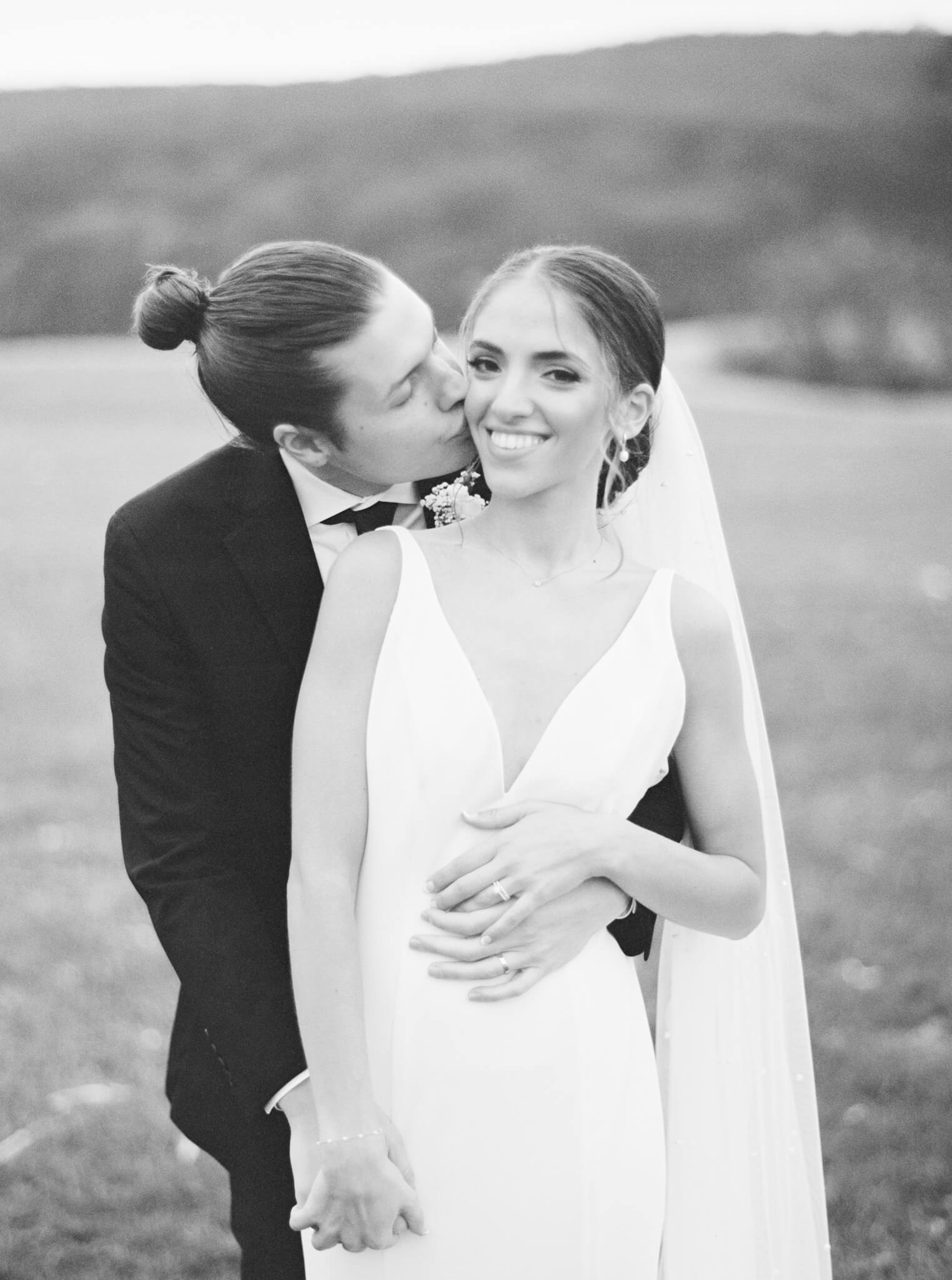 Black and white image of a groom kissing his bride on the cheek while she looks at the camera.