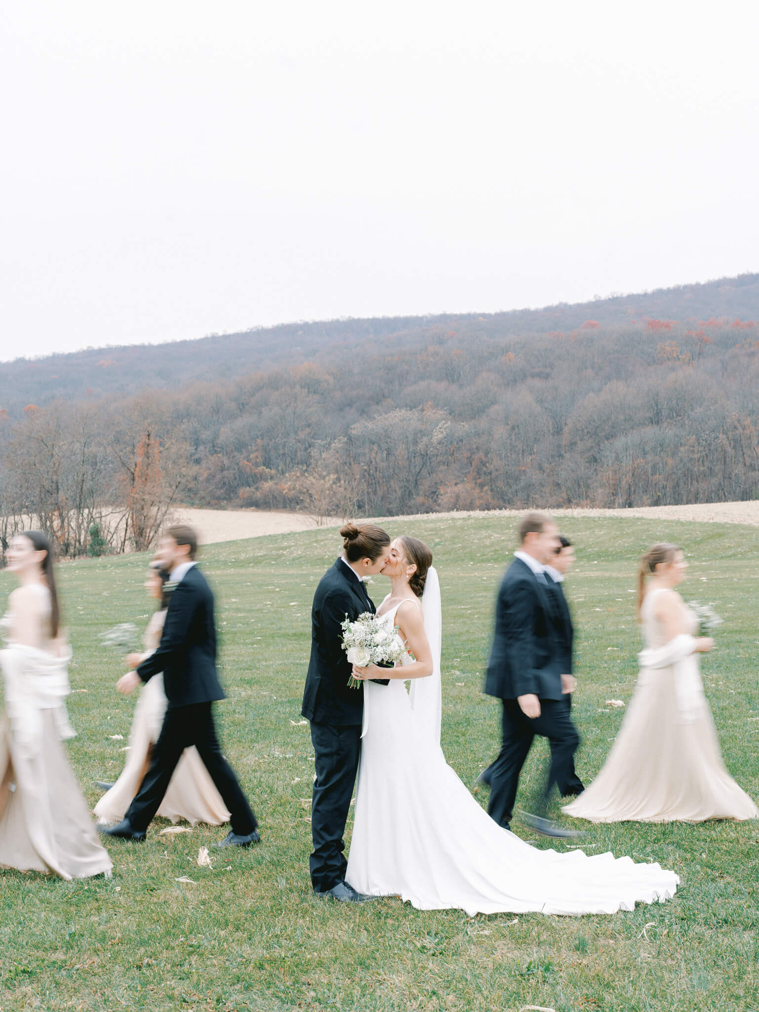 A bride and groom paused in time and kissing in front of a mountainous backdrop while their wedding party walks around them.
