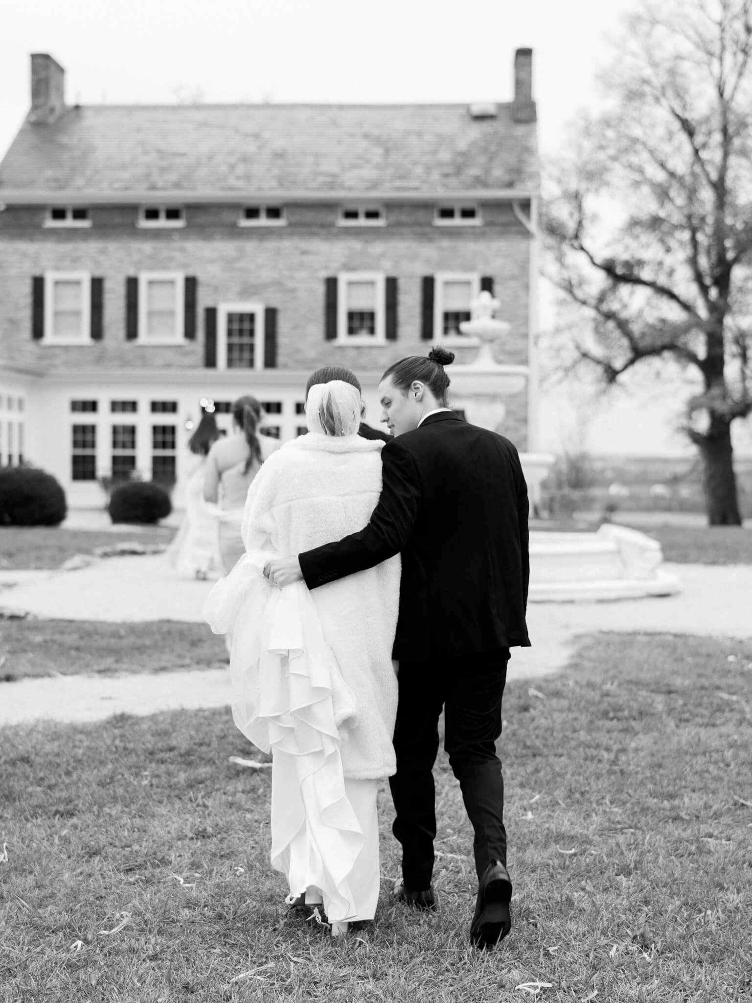 Black and white image of the bride and groom walking away towards their venue while the groom carries the back of the bride's gown.