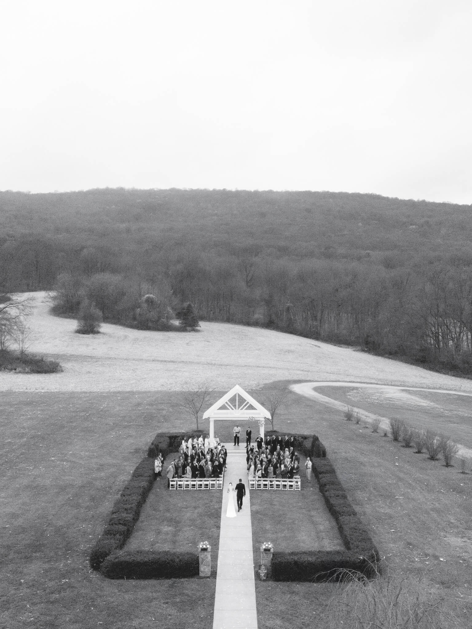 An black and white aerial view of the bride and her father walking down the aisle at a ceremony with the mountainous backdrop.