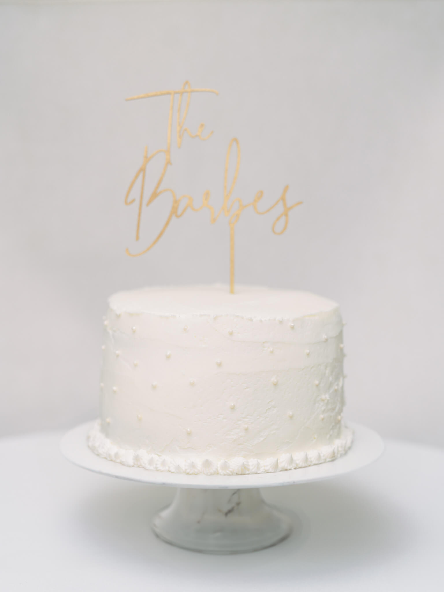 A one tier white wedding cake with pearls on a white marble cake stand with a gold worded topper.