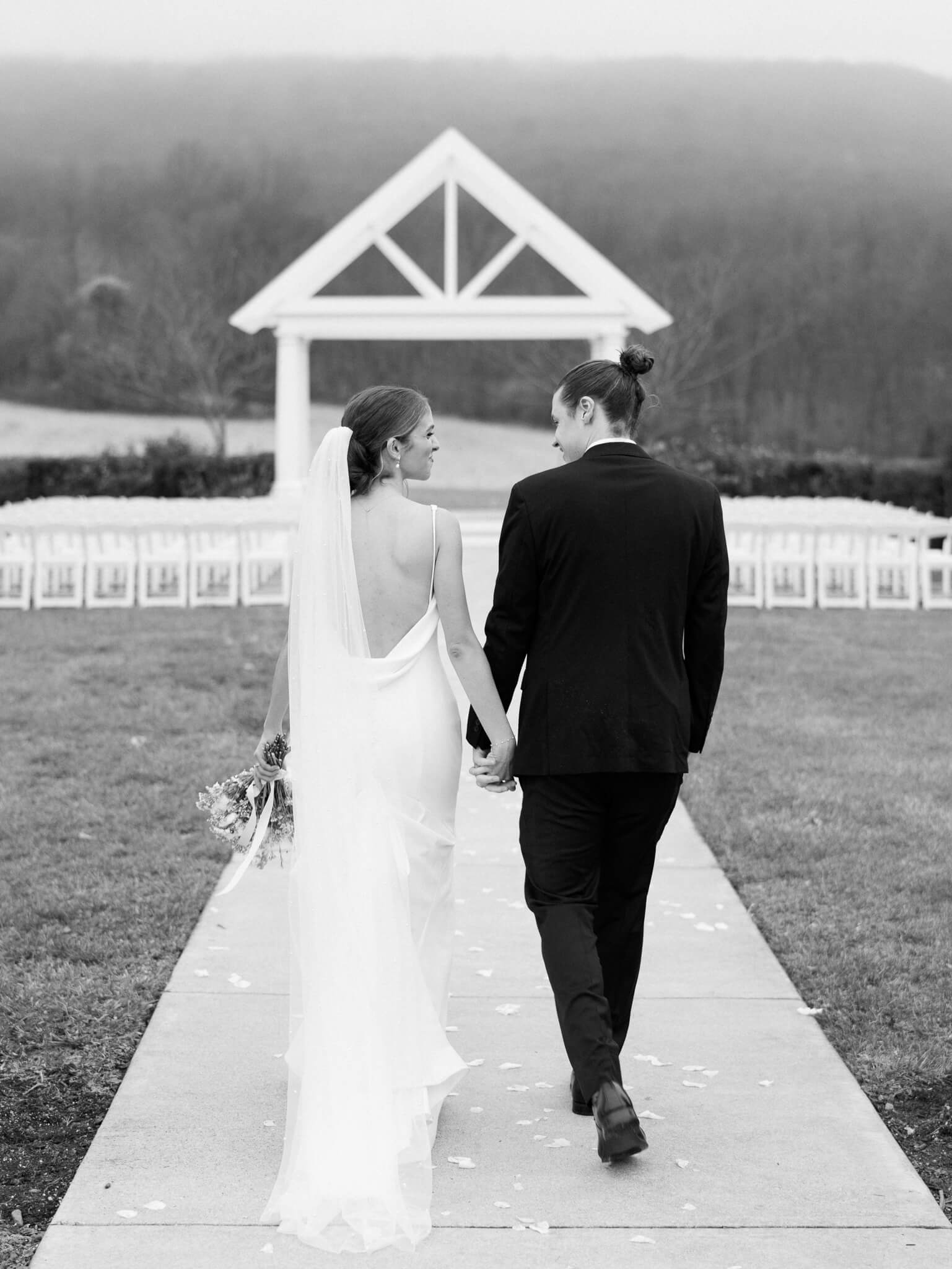A black and white of a bride and groom walking hand in hand away towards their ceremony space while looking at each other.