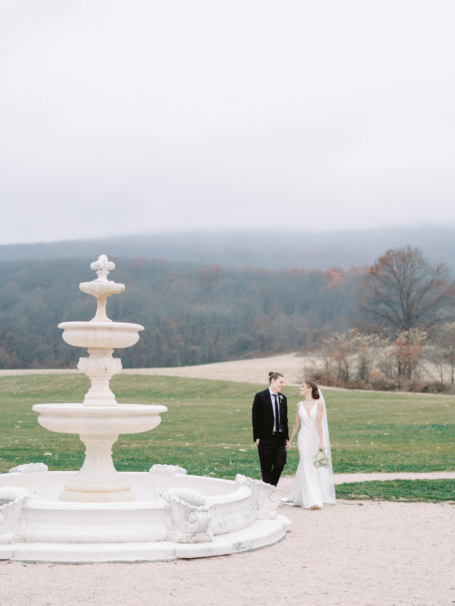 A bride and groom walking hand in hand next to Springfield Manor's fountain in front of a foggy, mountainous backdrop.
