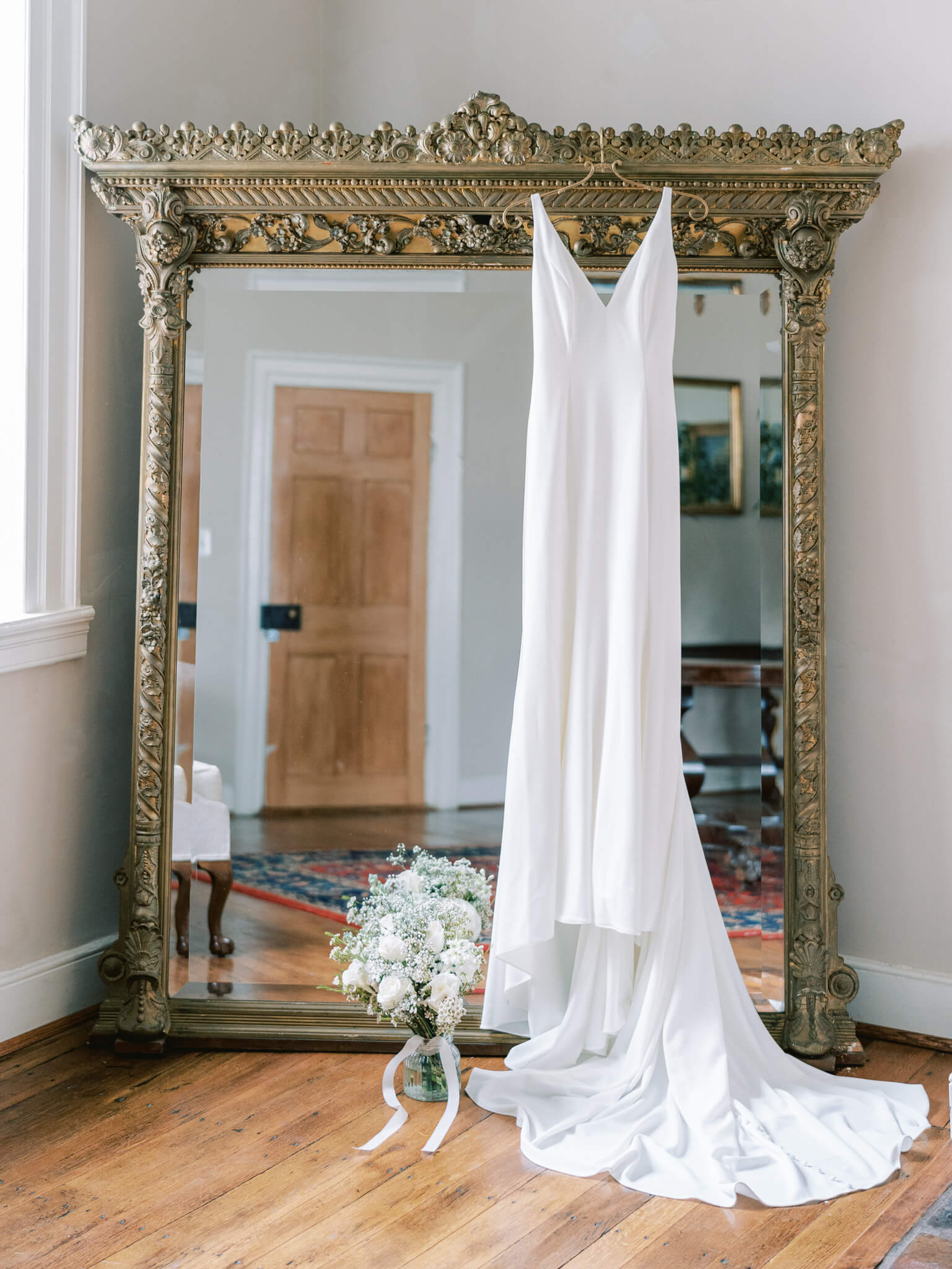 A sleek white wedding dress hanging from a large, ornate gold mirror with the bouquet and wedding shoes next to it.
